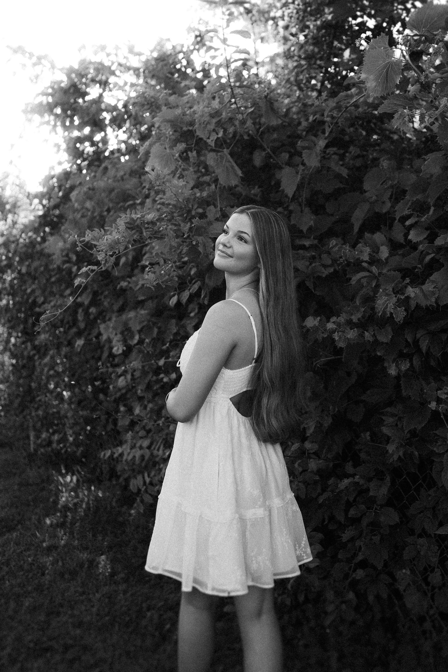 A black and white portrait against lush foliage wearing a flowing white sundress.