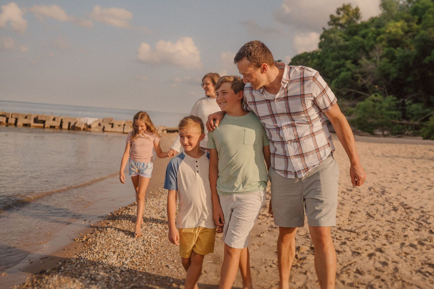 A family walks together along a sunny beach at sunset with a wooden pier visible in the background.