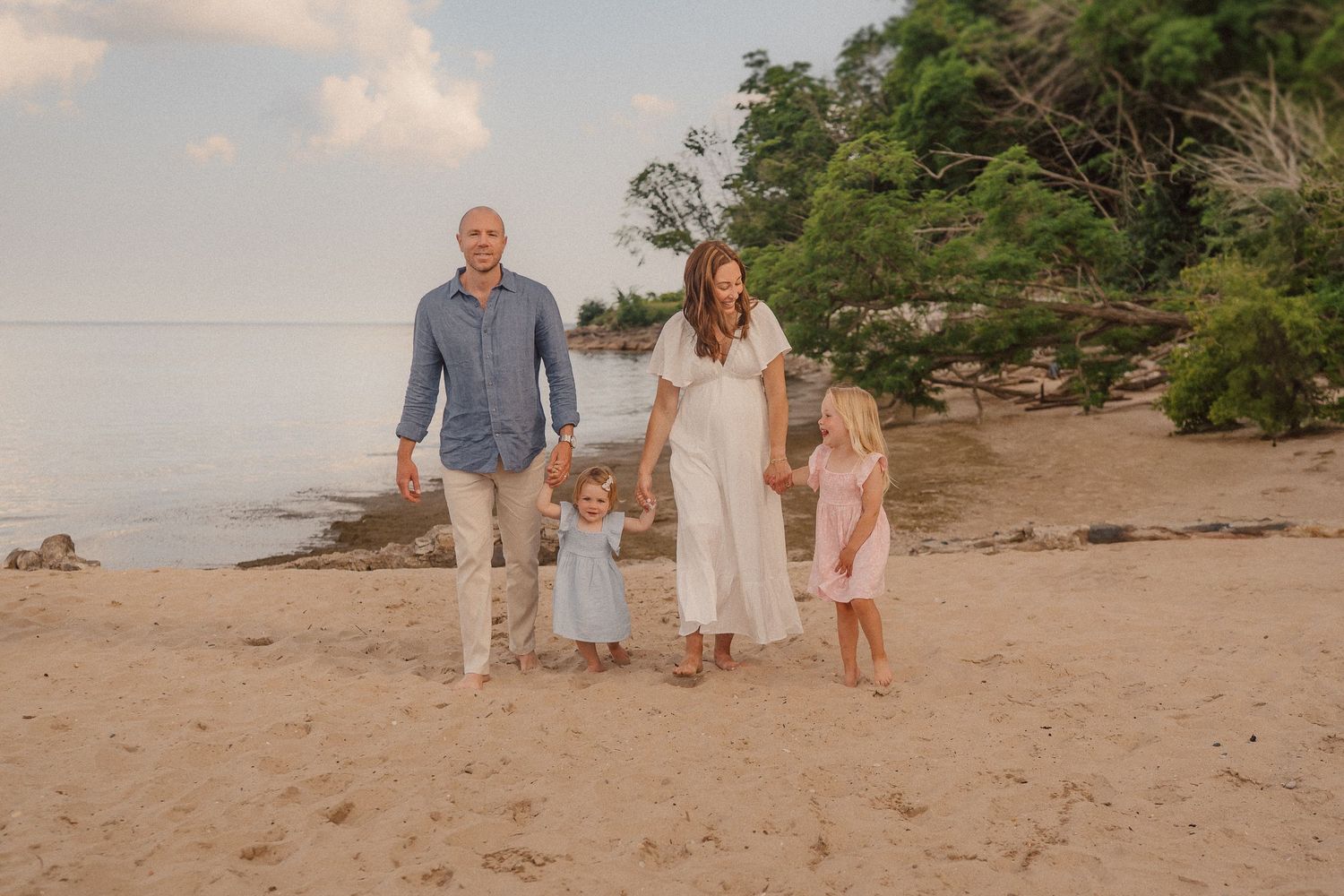 A family walks together along a sandy beach at sunset with trees and water in the background.