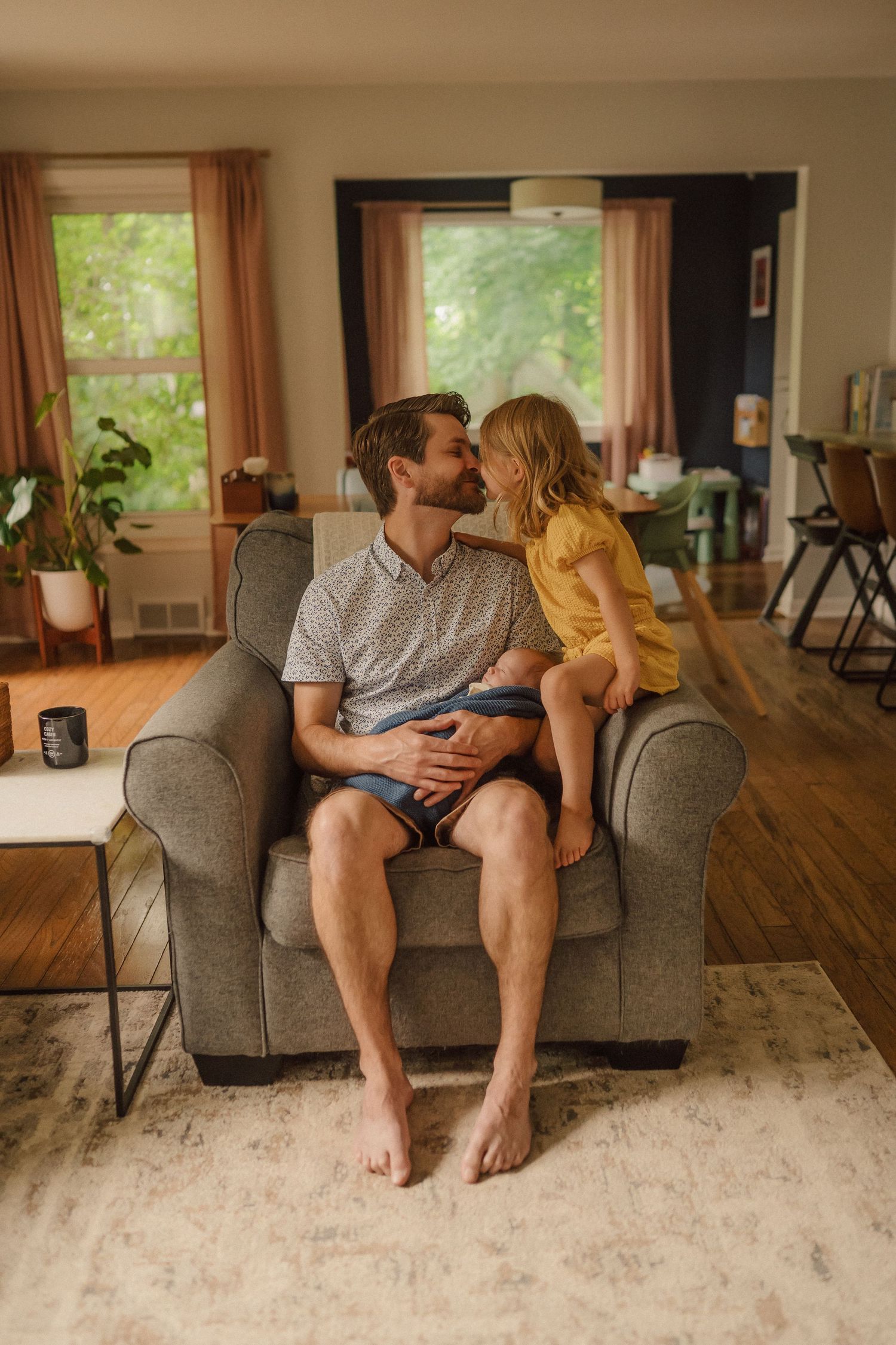 A tender moment shared on a gray armchair in a cozy living room with natural lighting from windows.