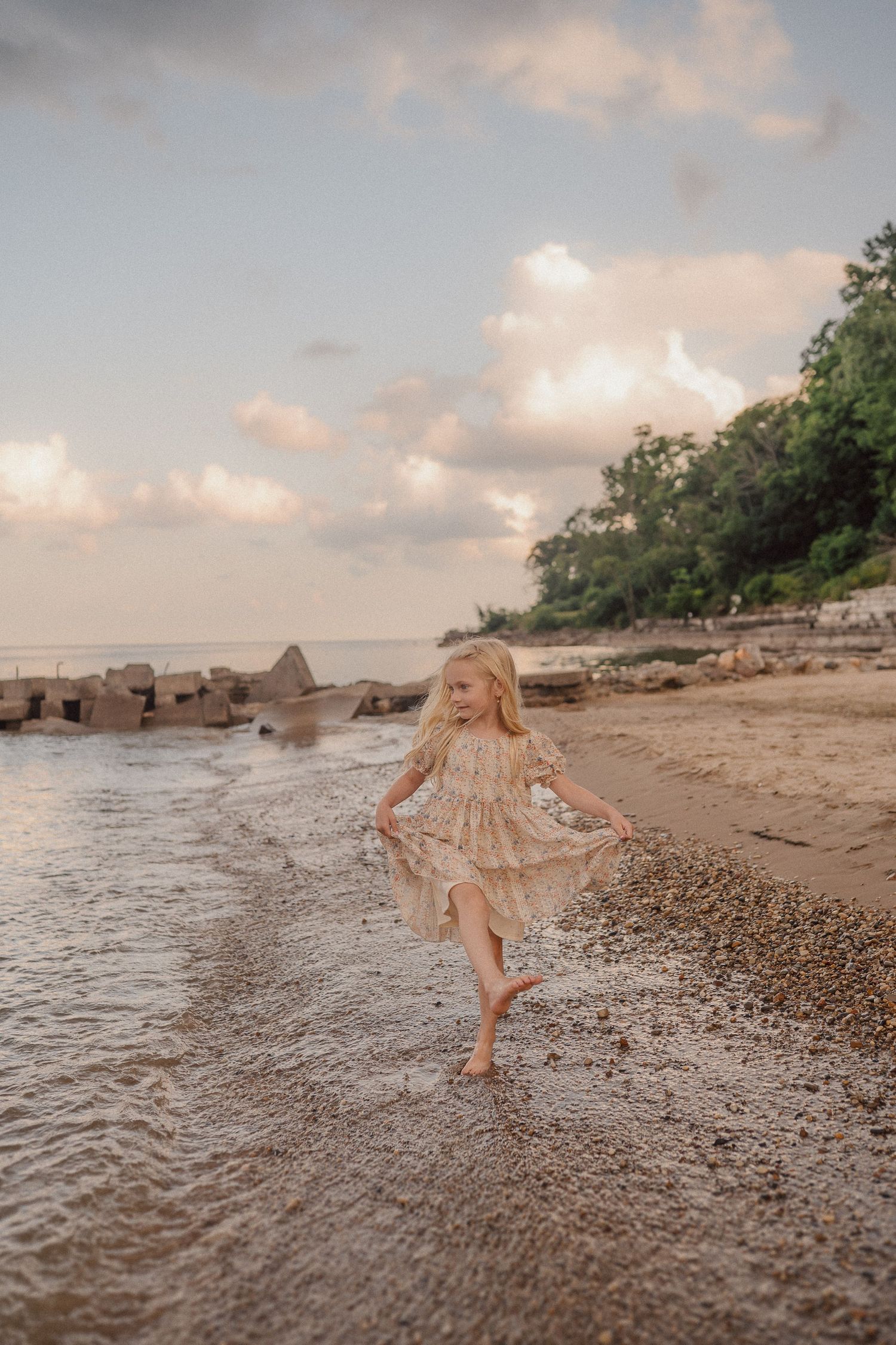 A young girl in a flowy dress dances at the water's edge during golden hour at a rocky beach.