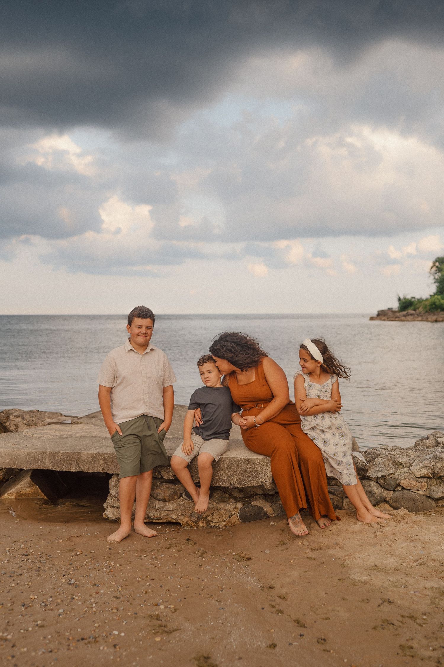 A family sits together on coastal rocks at the beach during sunset with dramatic cloudy skies above.