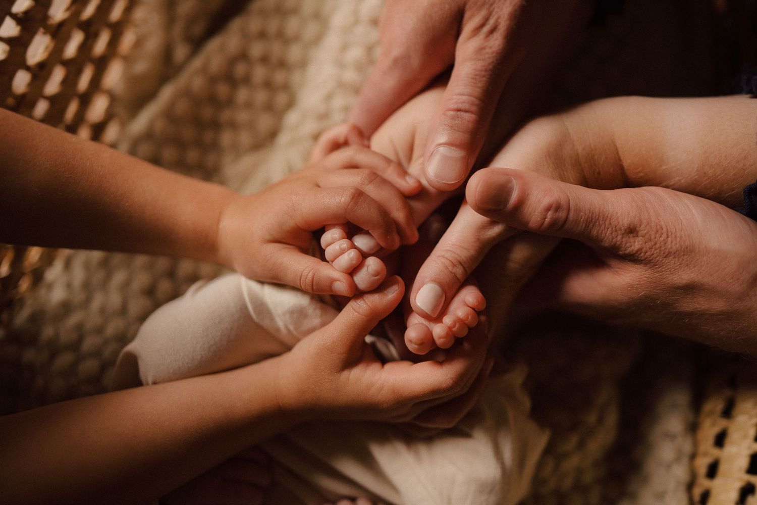 Multiple hands tenderly holding and touching a baby's tiny fingers in warm lighting.