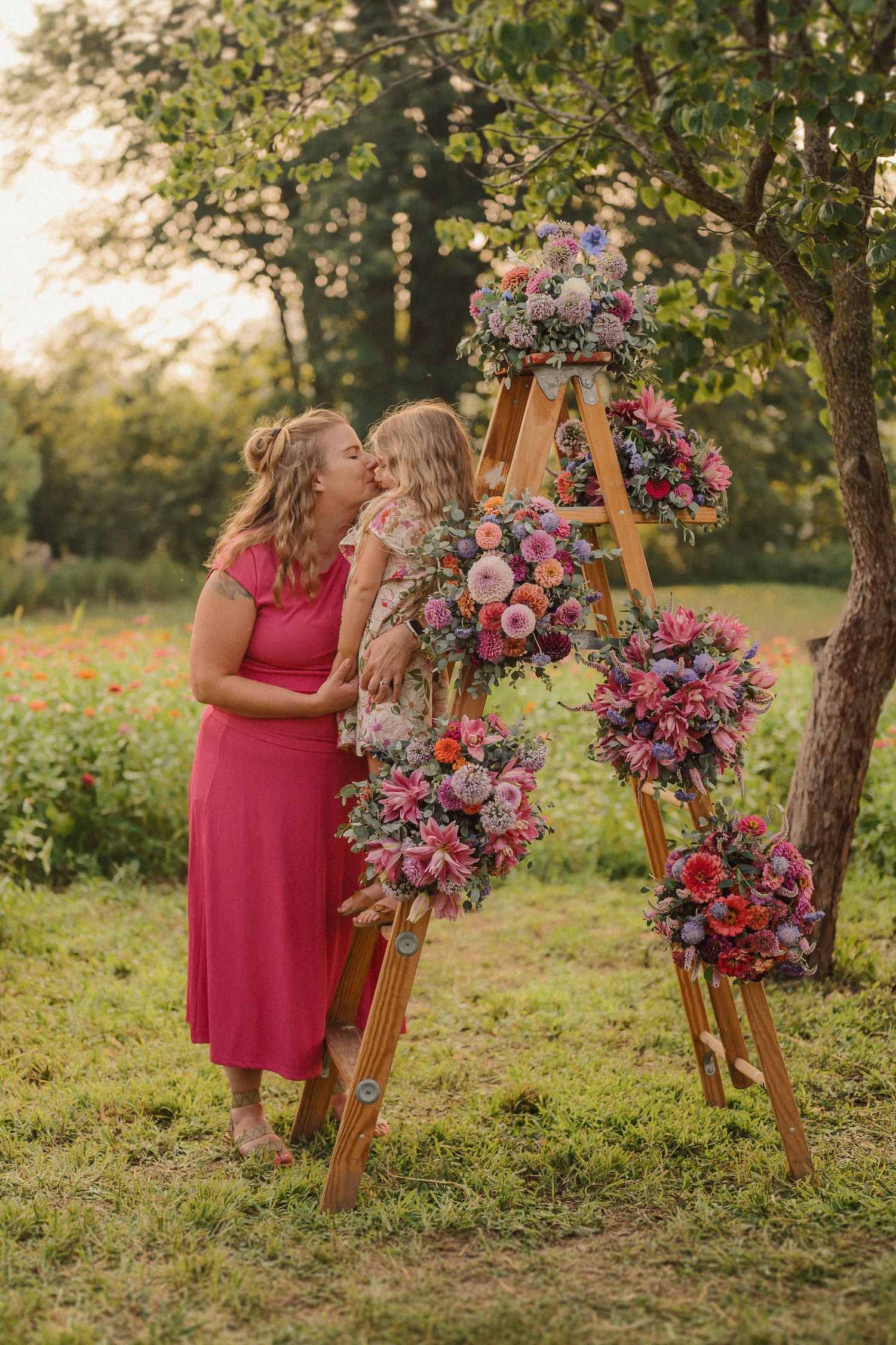 In a grassy garden, a person in a pink dress stands beside a wooden arch adorned with pink and red flowers.