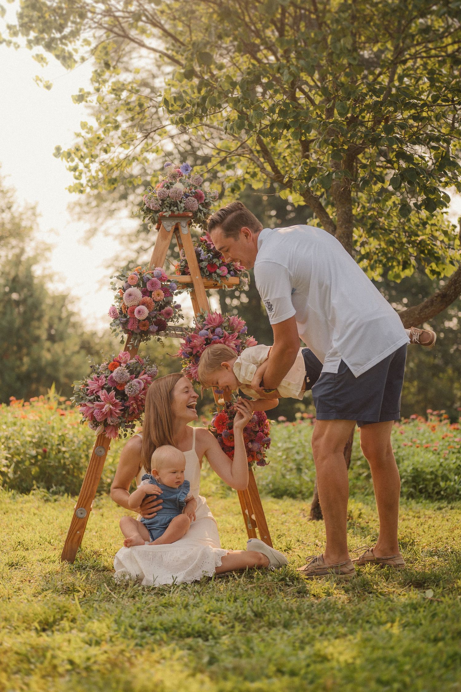 Family playing and laughing together on grass with pink flower crown and summer sunlight filtering through trees.