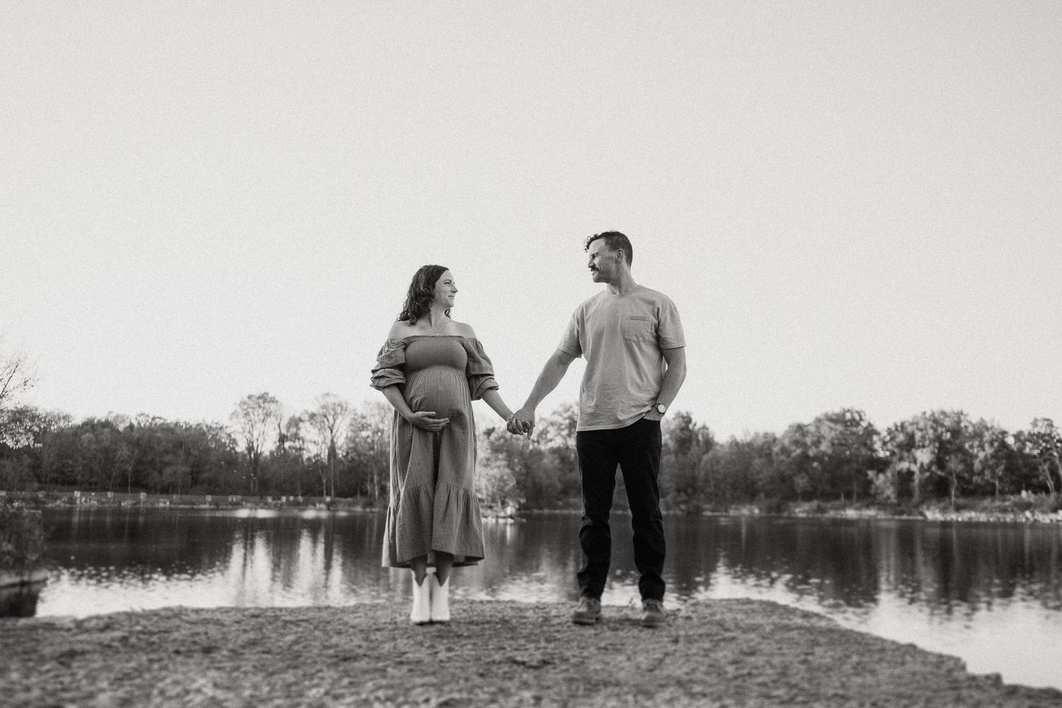Black and white photo of couple holding hands by serene lakeside at sunset.
