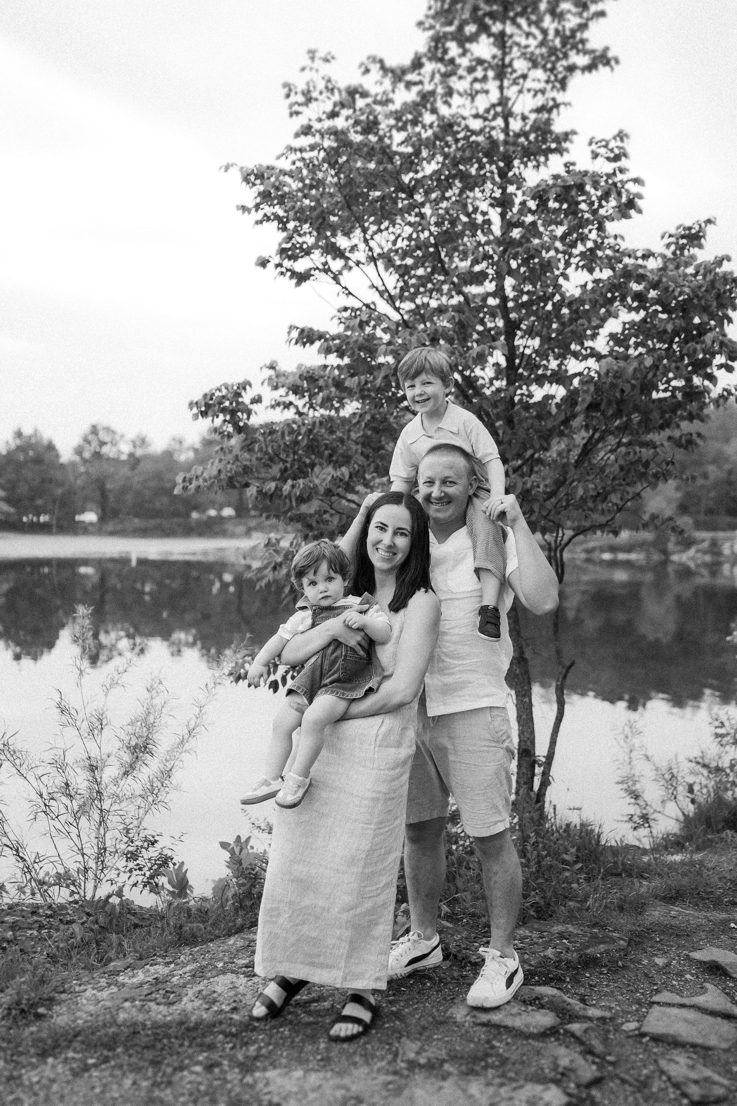 A family stands together by a serene lake on a summer day, with a leafy tree creating a natural backdrop in black and white.