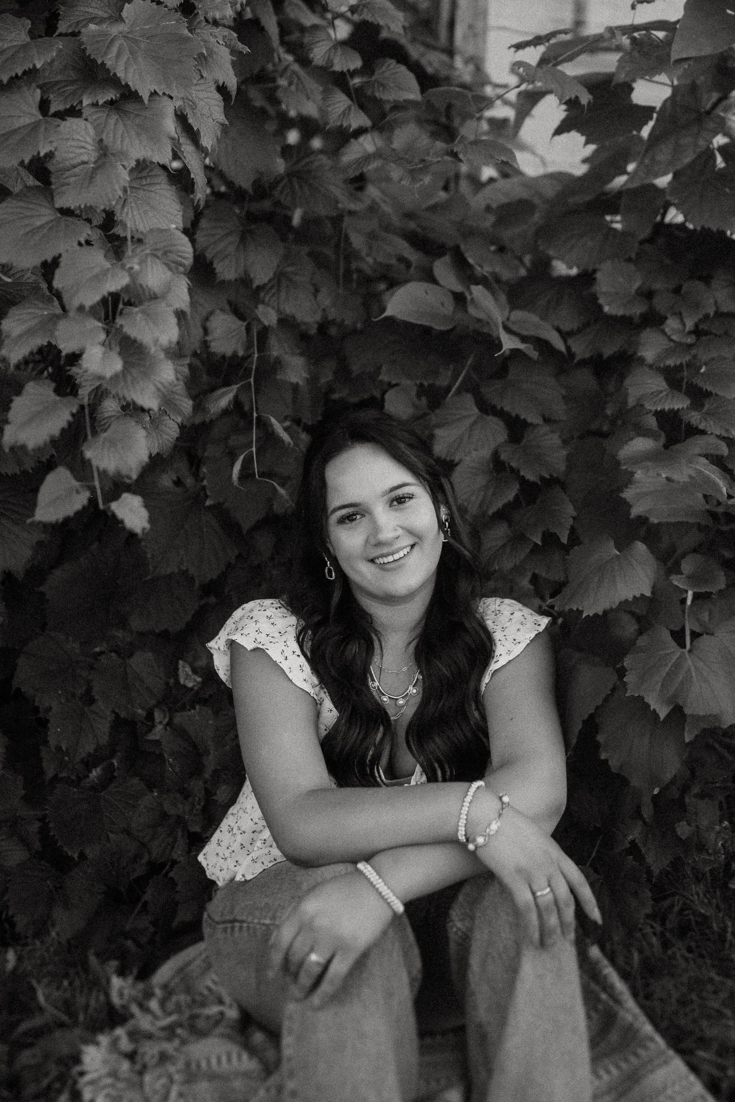 Black and white portrait of a smiling person sitting among leafy vines wearing a lace top and jeans.