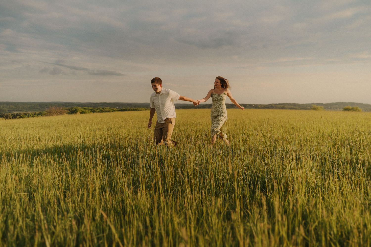 Couple walking hand in hand through golden wheat field at sunset with cloudy sky overhead.