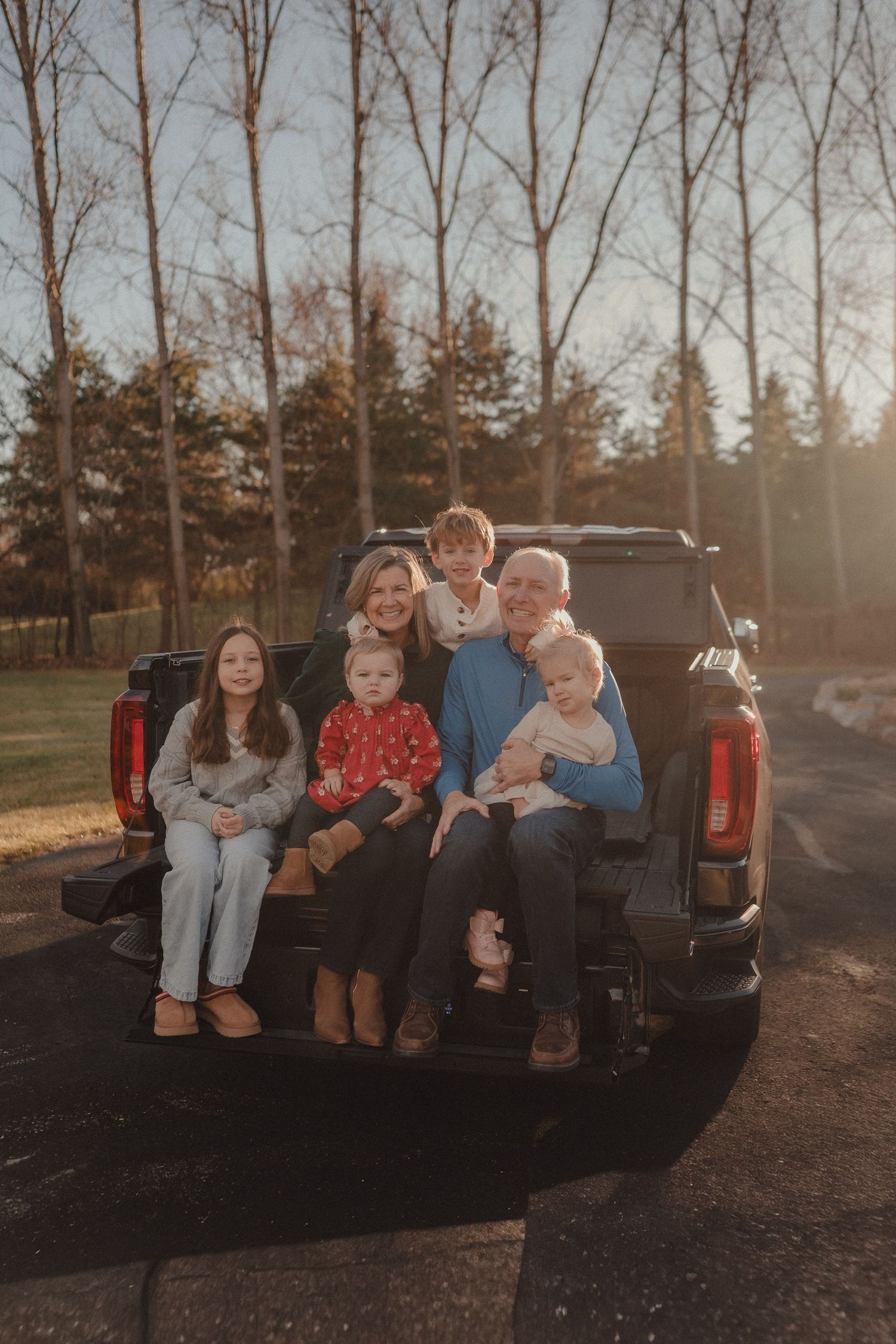 A family sits together on the back of a pickup truck at sunset surrounded by bare trees.