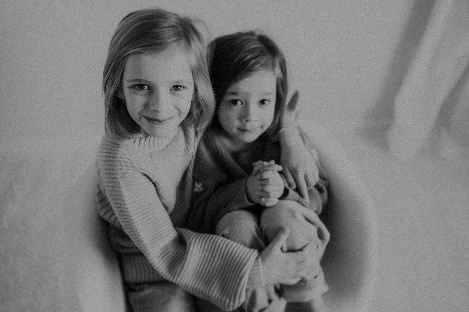 Two children in sweaters posing together on a modern chair with a stuffed toy.