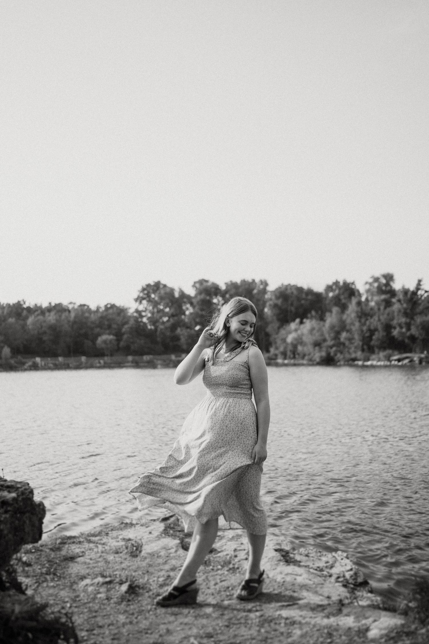 Black and white photo of a woman in dress standing by water with wind-swept hair and dress.