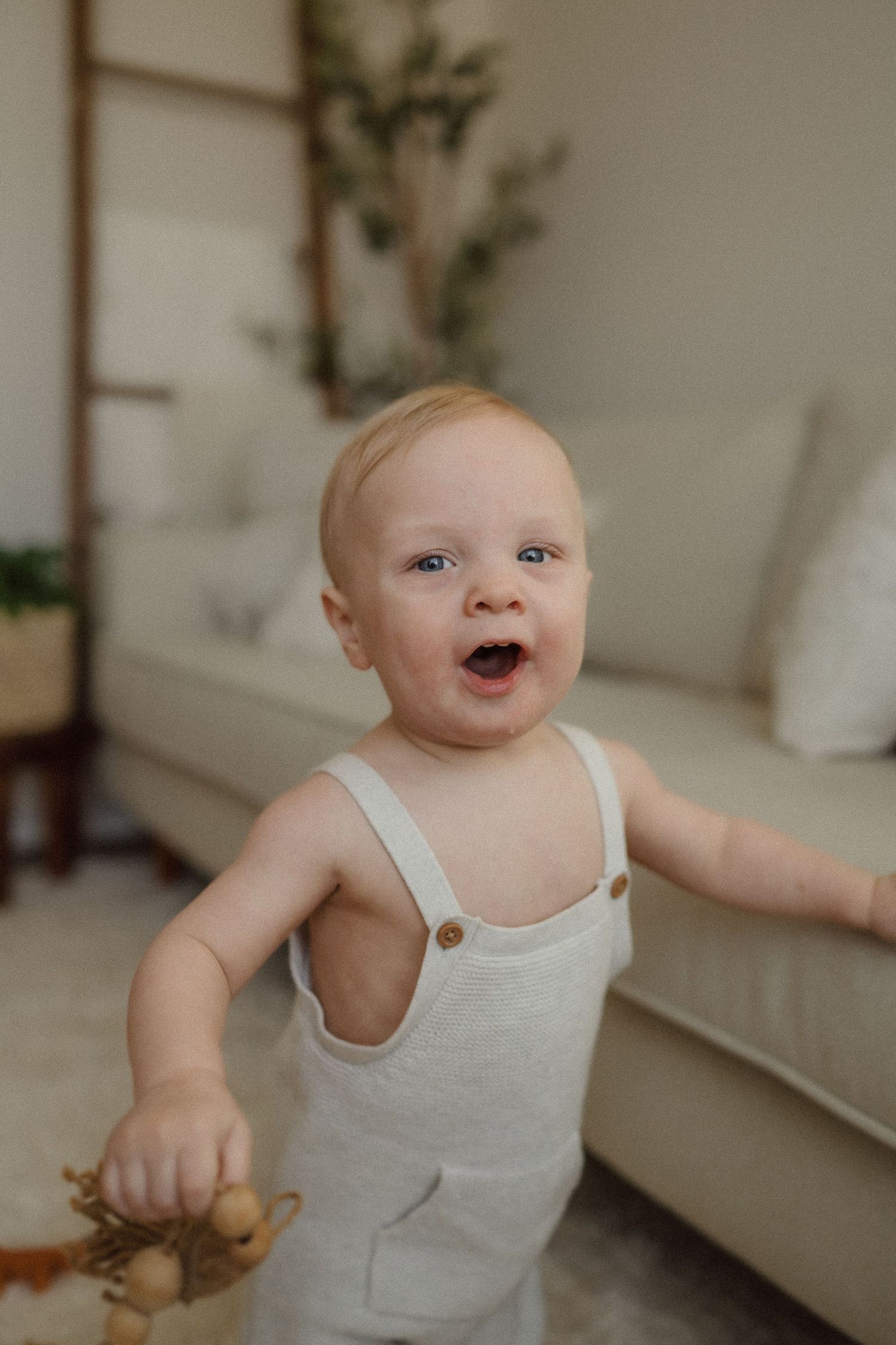 A joyful baby in white overalls plays and smiles in a minimalist living room setting.
