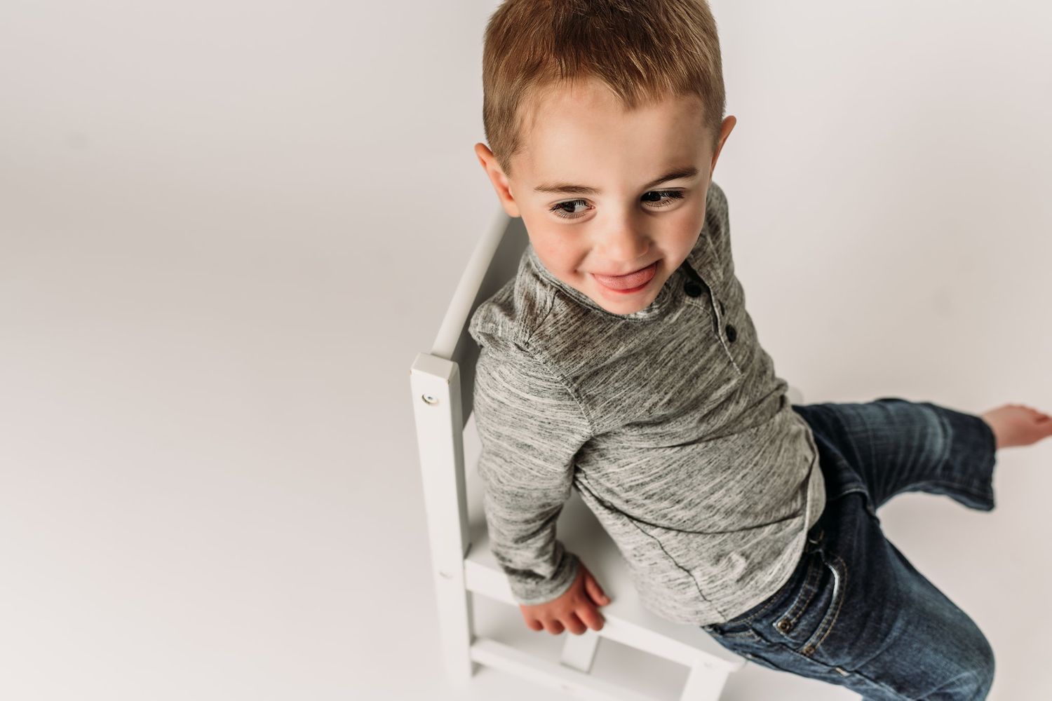 Child climbs on white furniture in casual attire during studio photoshoot.