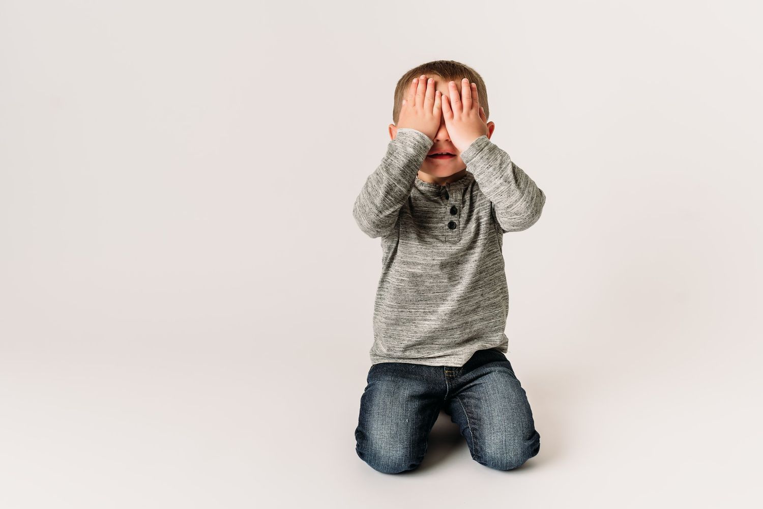 Playful pose covering eyes with hands while wearing a gray sweater on white background.