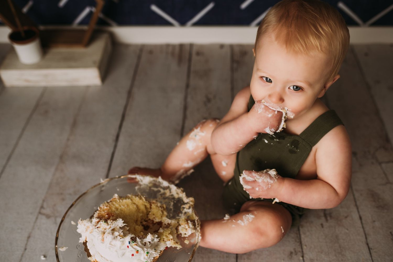 Baby in green outfit explores cake on wooden floor against geometric patterned wall.
