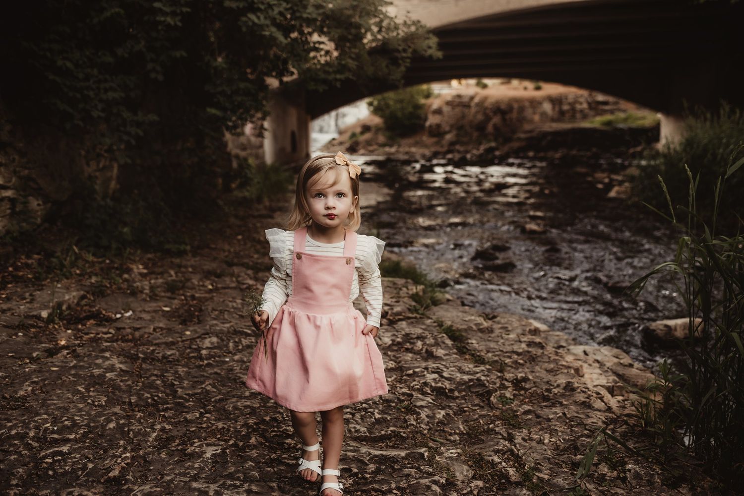 A child in a pink dress stands by a stone bridge over a creek.
