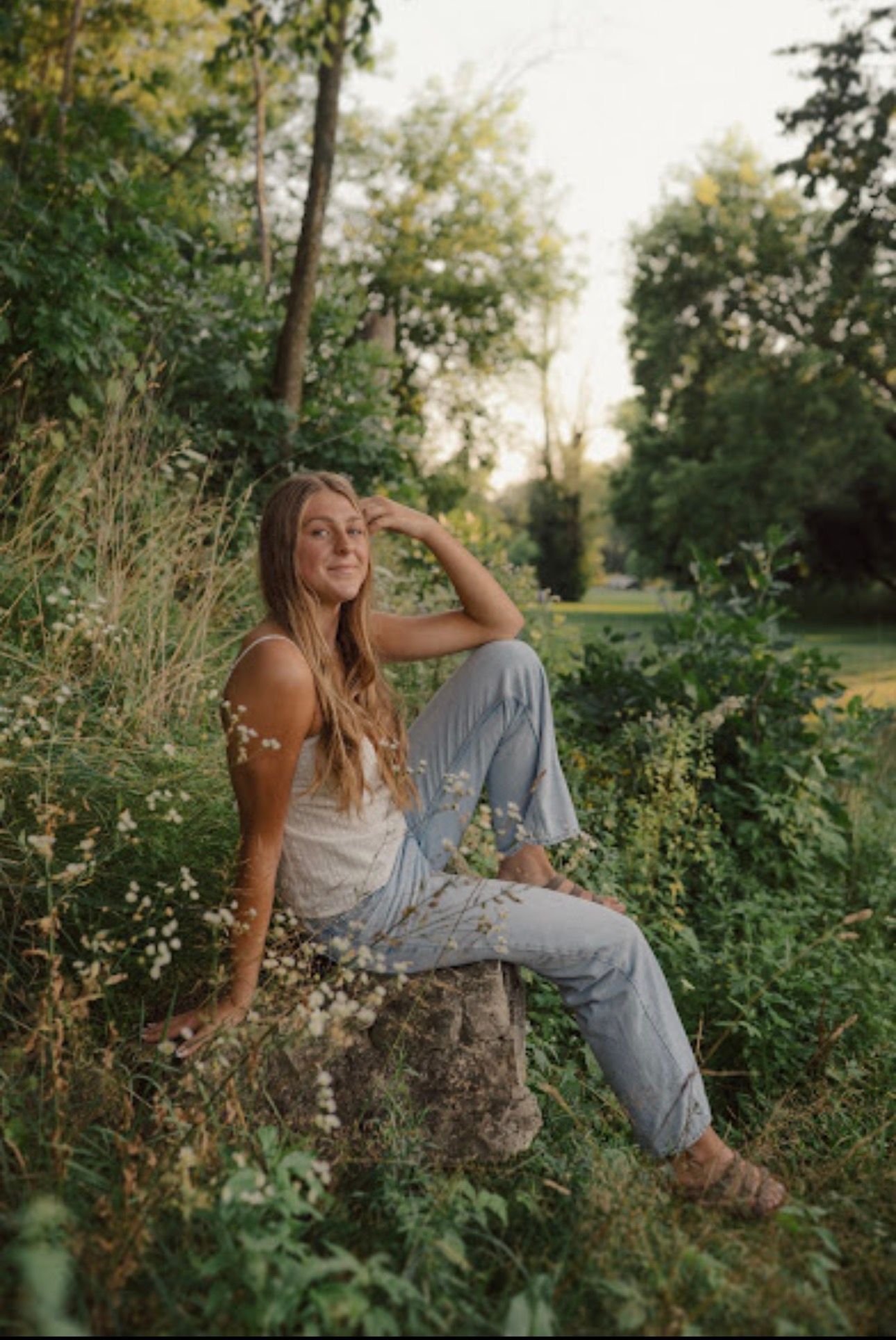 Candid outdoor portrait of someone sitting on a rock in denim, surrounded by greenery at golden hour.