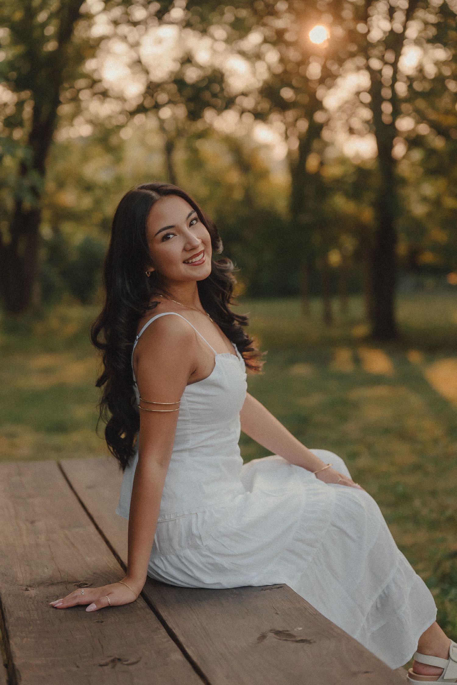 Young woman in white dress sitting on wooden dock smiling in a sunlit park at golden hour.