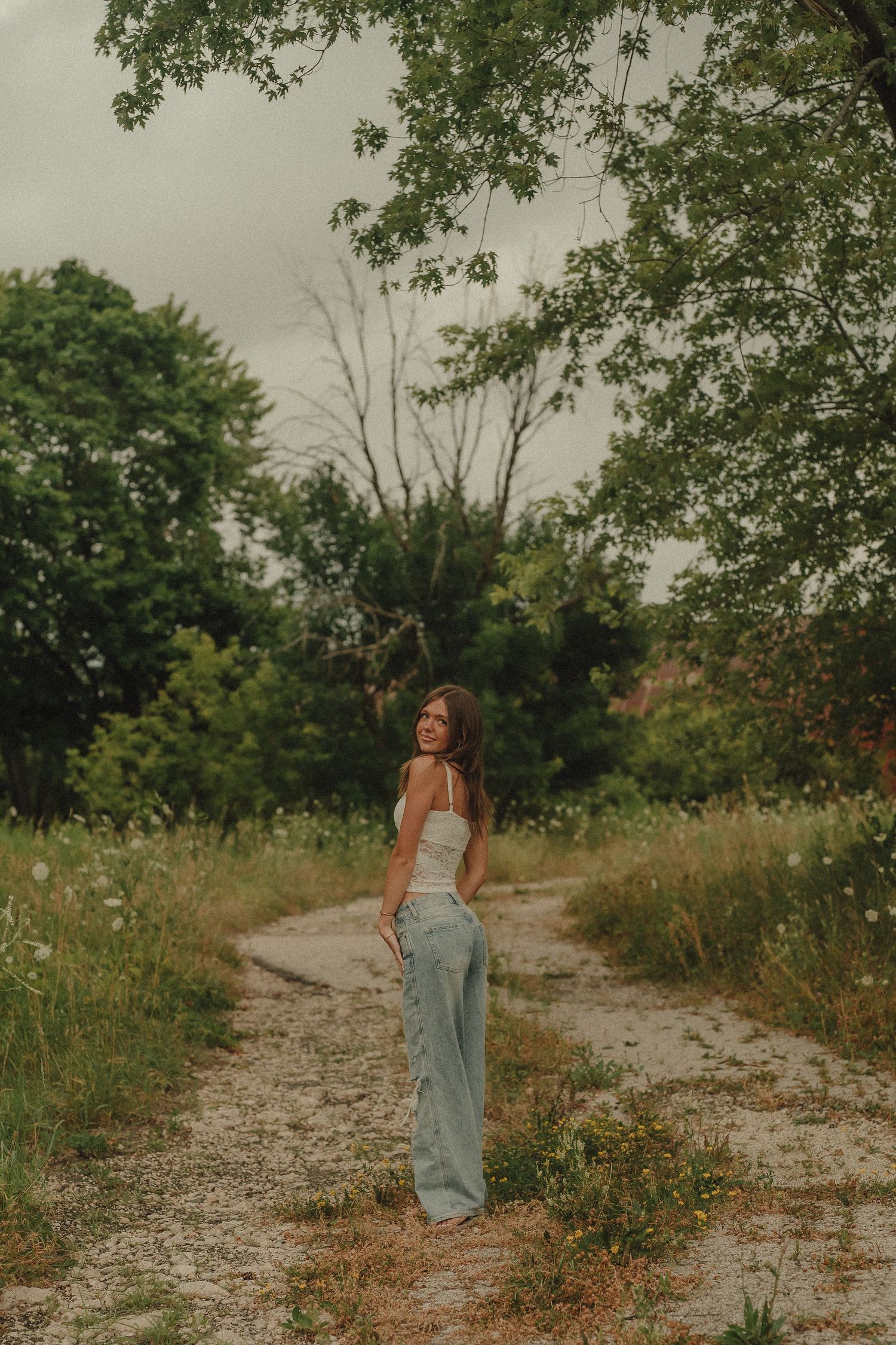 Young woman in white top and jeans standing on dirt path surrounded by lush green trees.