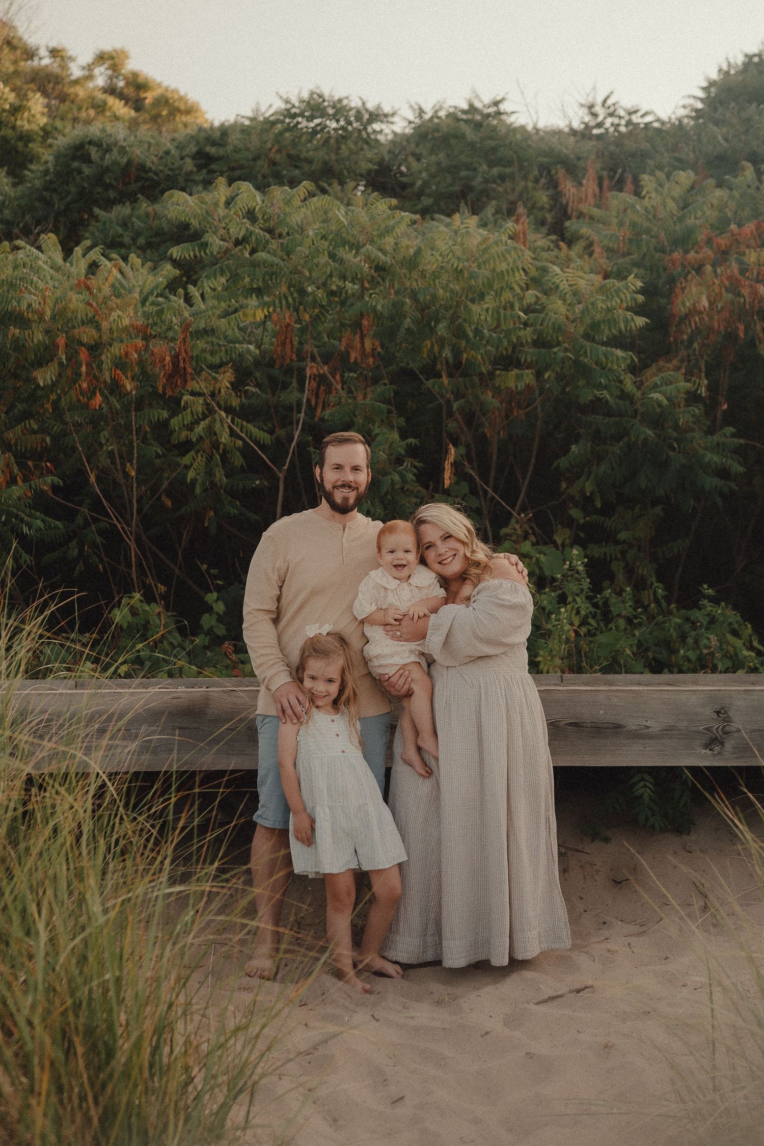 Family of four poses on sandy beach path surrounded by lush green foliage at sunset.