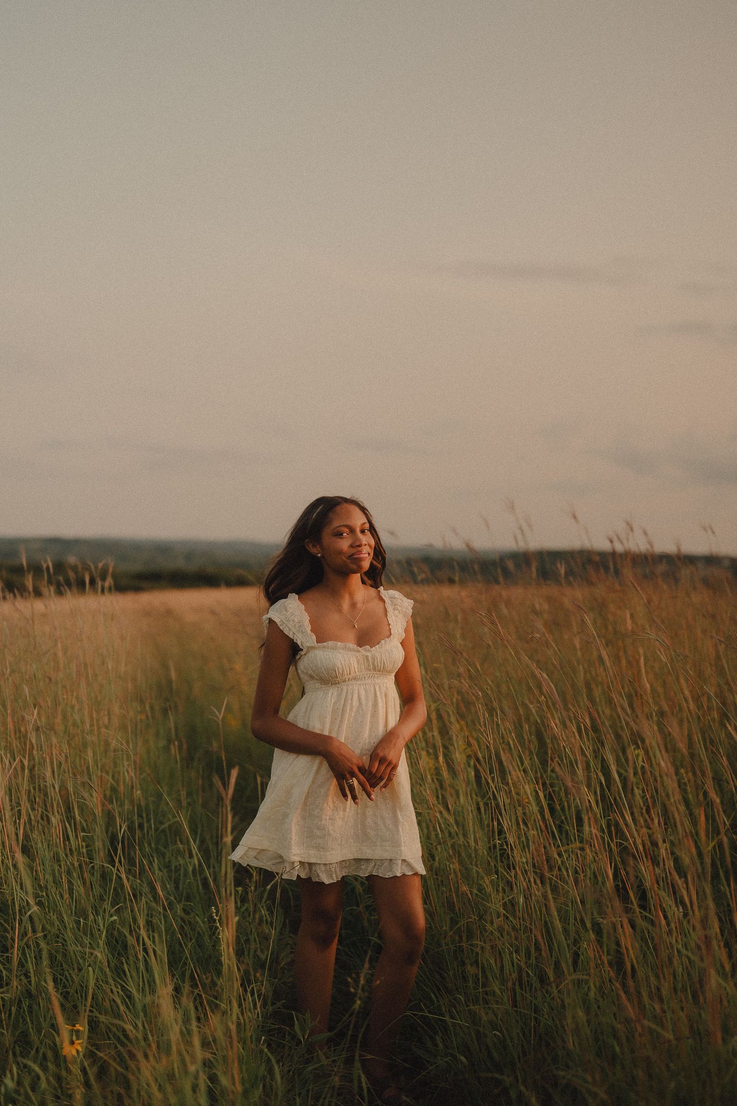 Young woman in white lace dress standing in tall golden grass field during warm sunset.