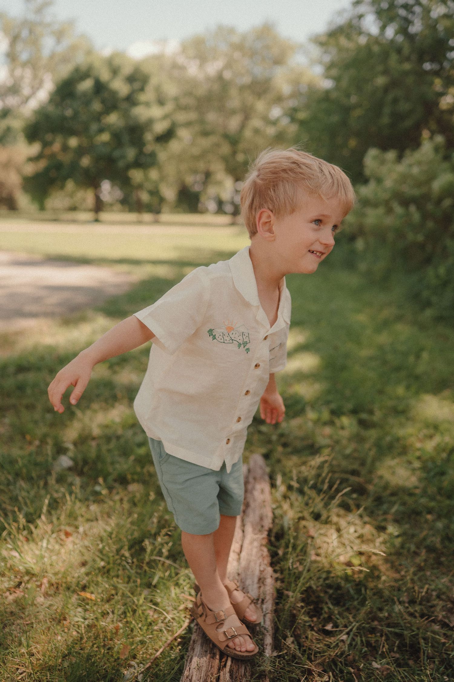 A cheerful toddler in a cream linen shirt and teal shorts balances on a log in a sunny park.