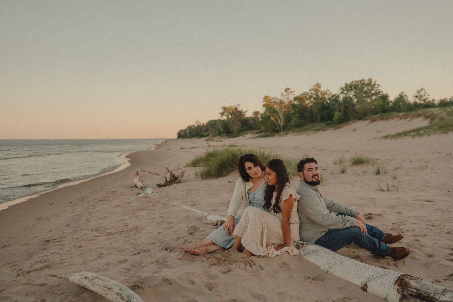 Three friends sit together on a sandy beach at sunset with dunes and trees in the background.