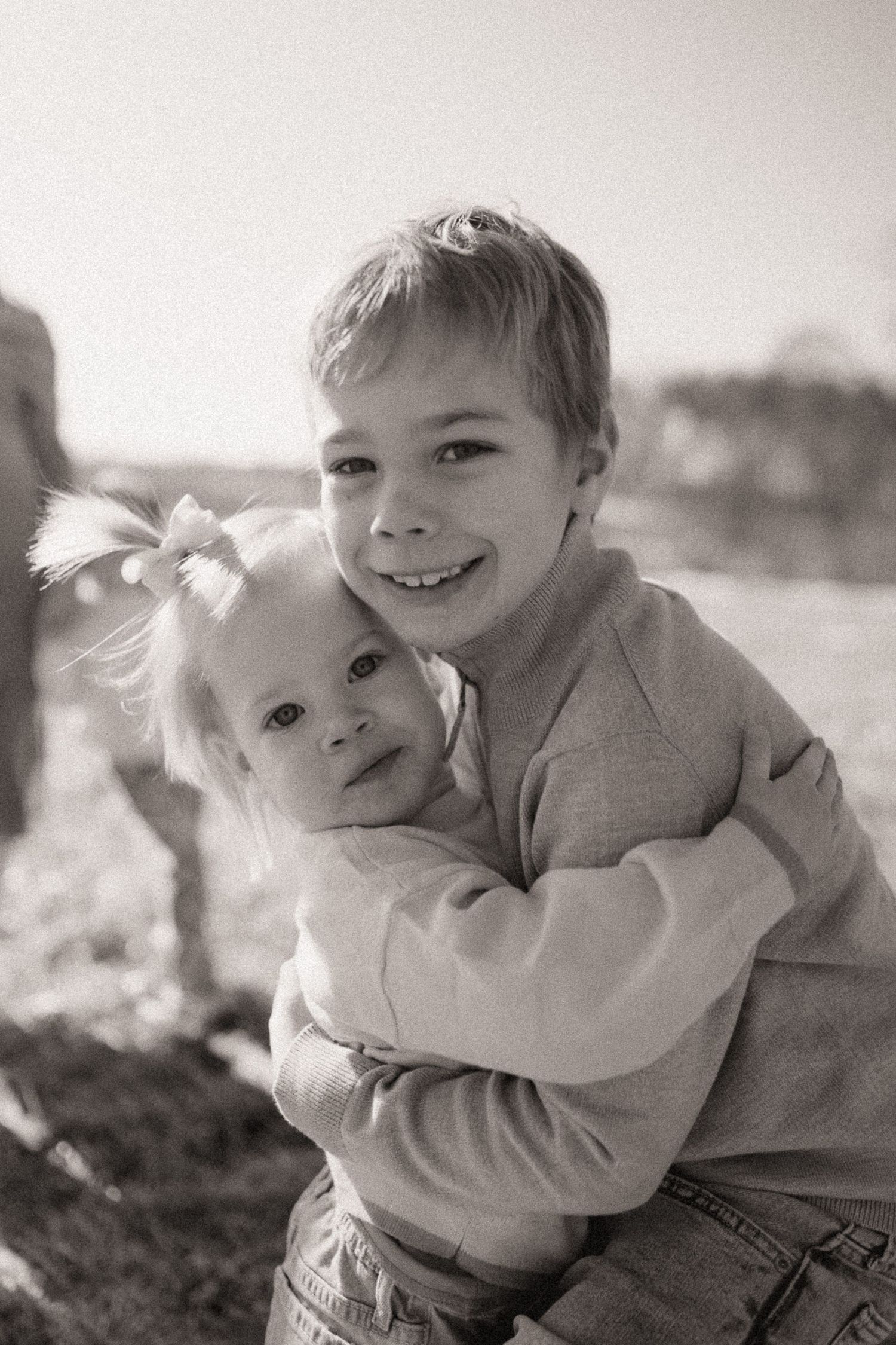Two young children in light colored sweaters share a playful embrace outdoors.