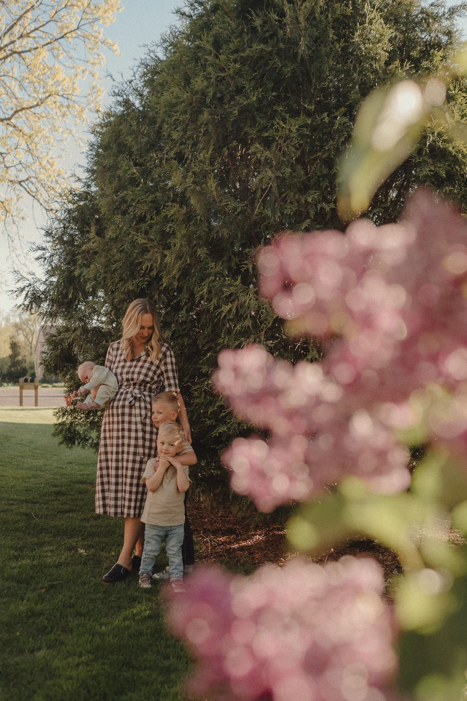 A smiling mother in a plaid dress holds her baby near lilac bushes while her toddler stands beside her.