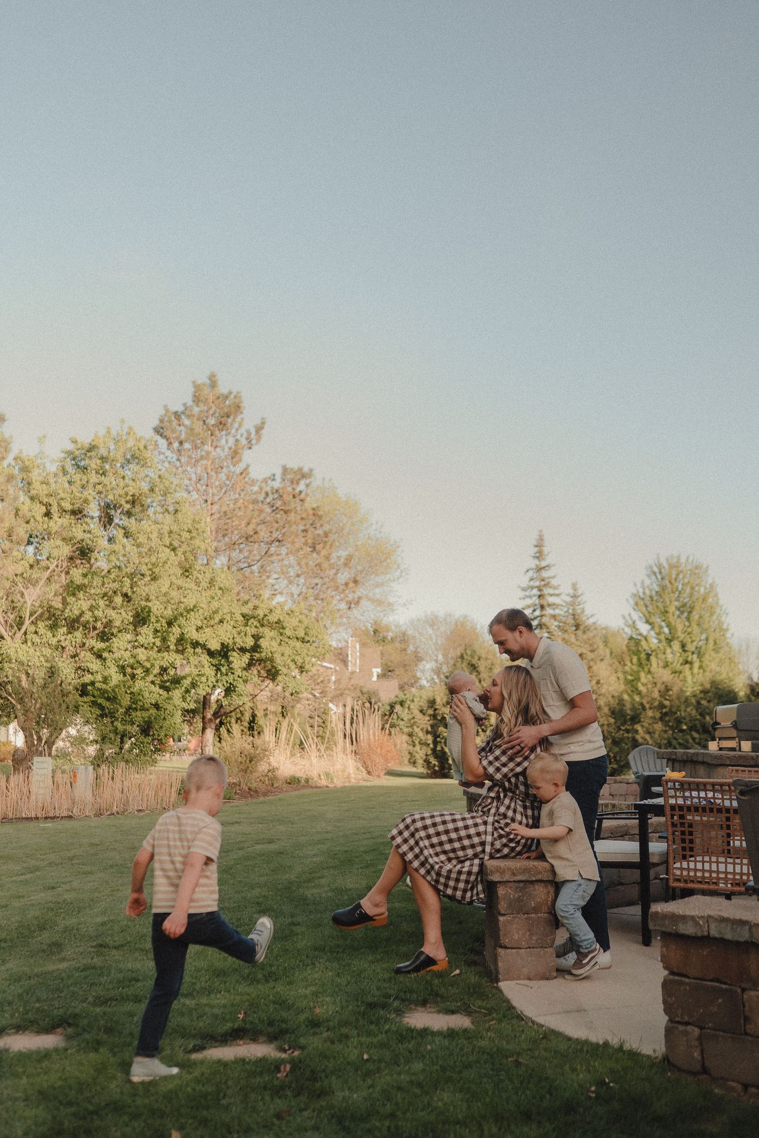 A family enjoys a sunny backyard gathering, with children playing on green grass near a stone patio.