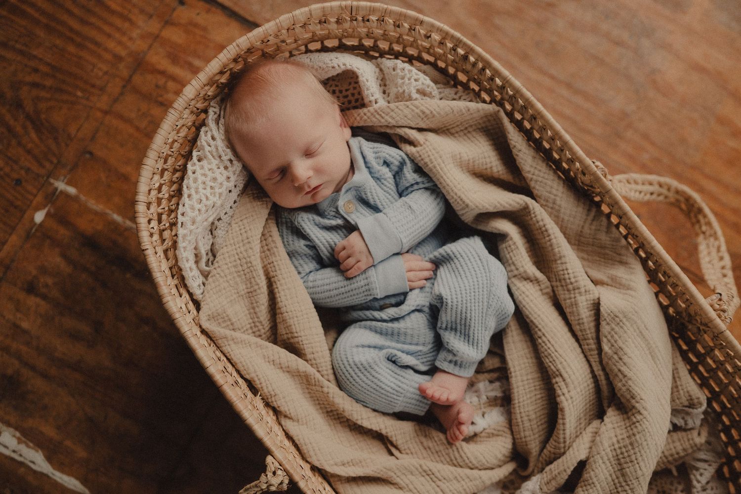 Newborn baby sleeping peacefully in a wicker Moses basket with a beige knit blanket, wearing a blue ribbed outfit.