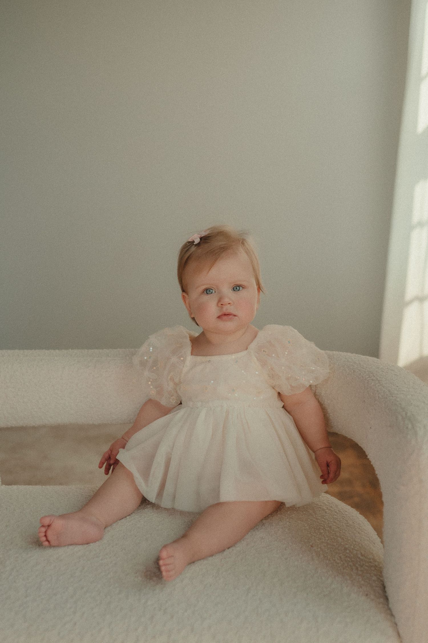 Adorable baby girl in a white lace puff sleeve dress sits on a cream chair during her first birthday photos.