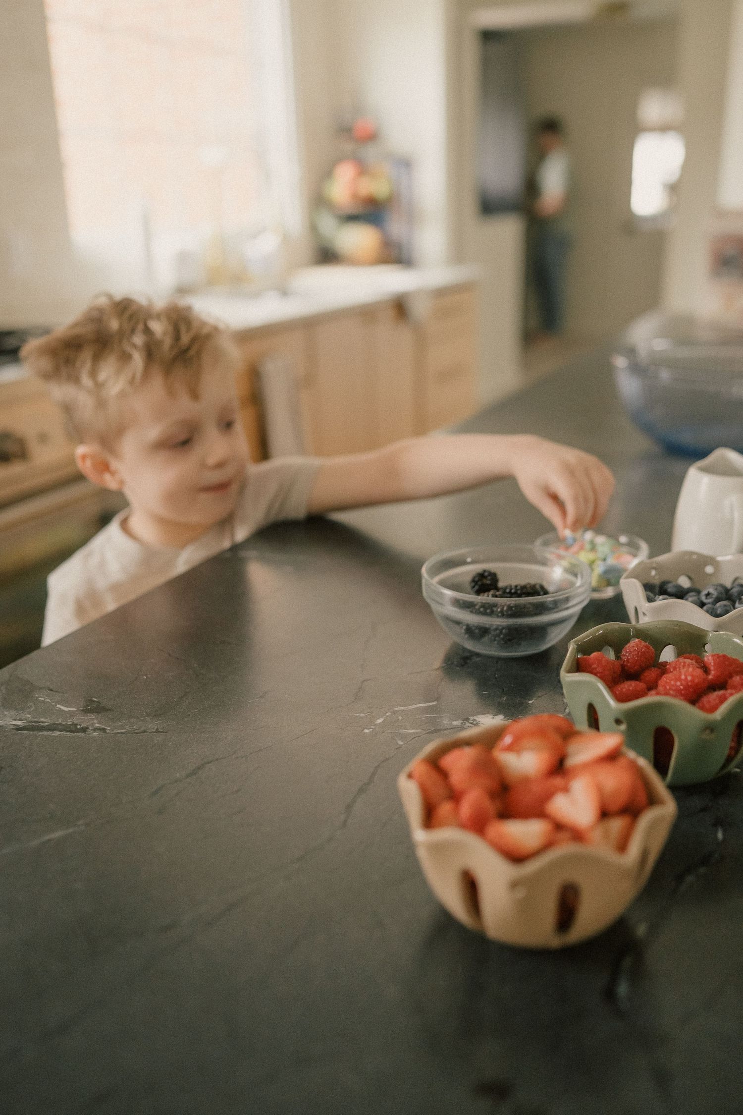 Young curly-haired boy reaches for berries in bowls on a dark stone kitchen counter.