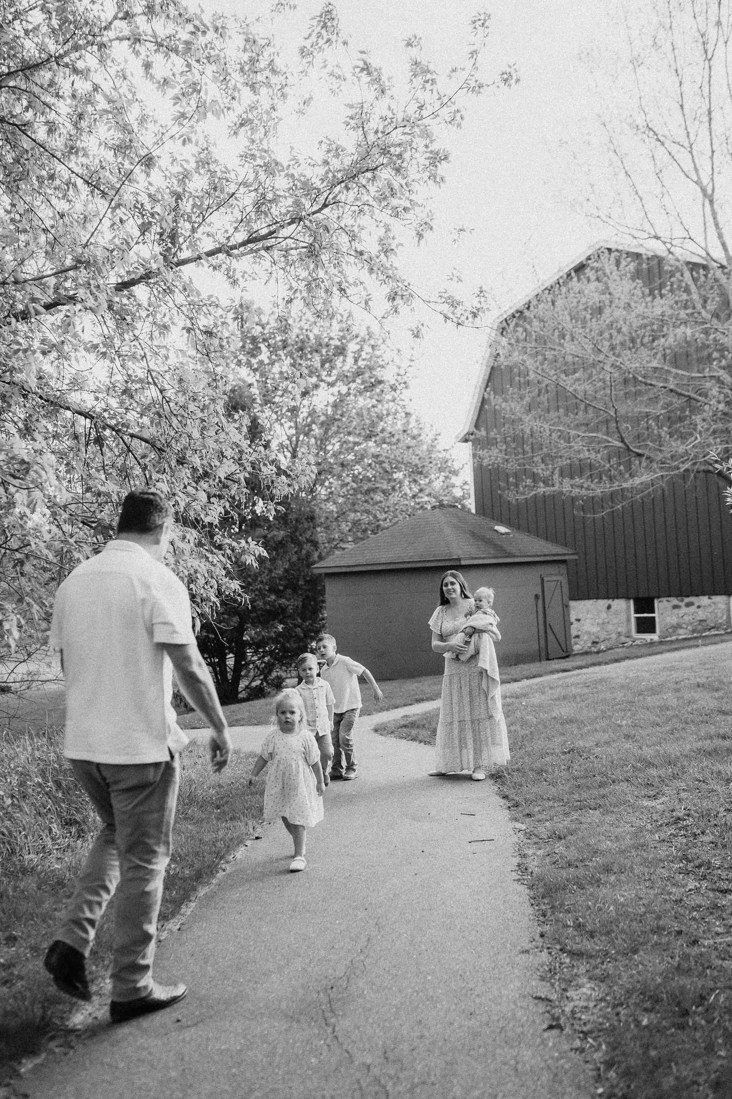 Black and white photo of a family walking on a path near a barn, with children running toward their father.