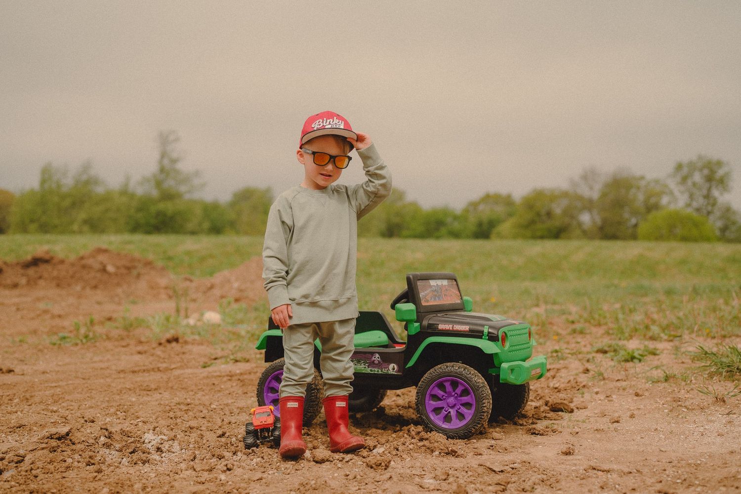 Young boy in red boots and cap poses with a green toy Jeep in a muddy field on a cloudy day.