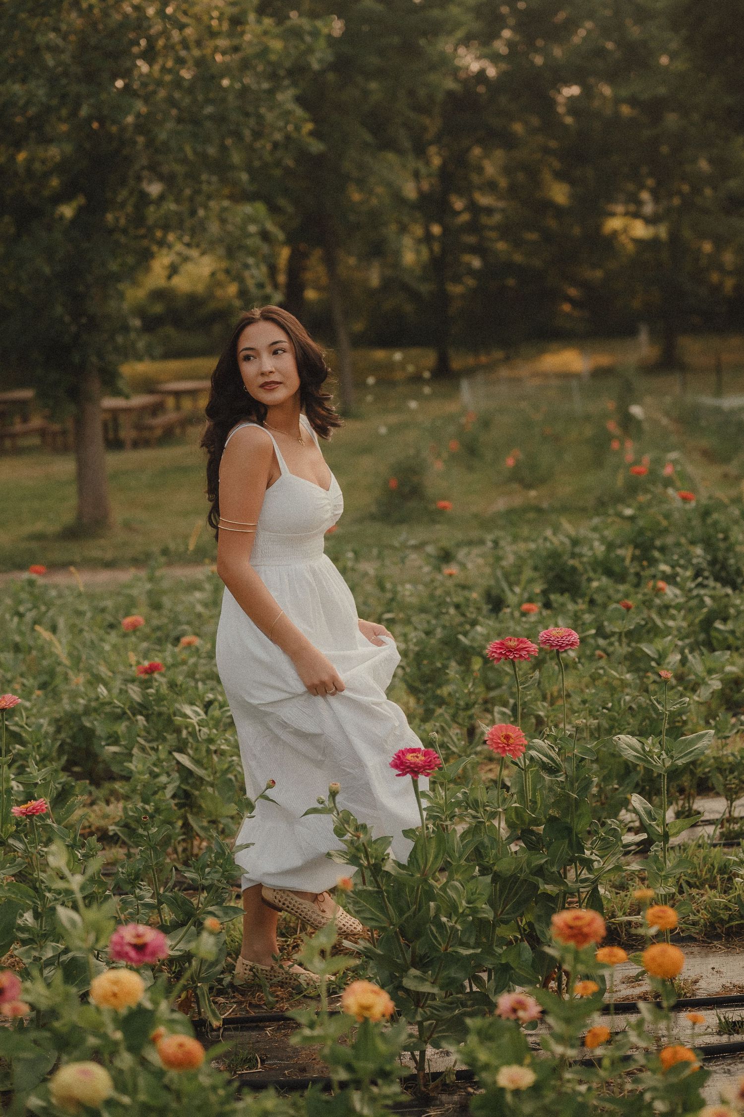 Woman in white dress standing among colorful zinnia flowers in a lush garden at golden hour.