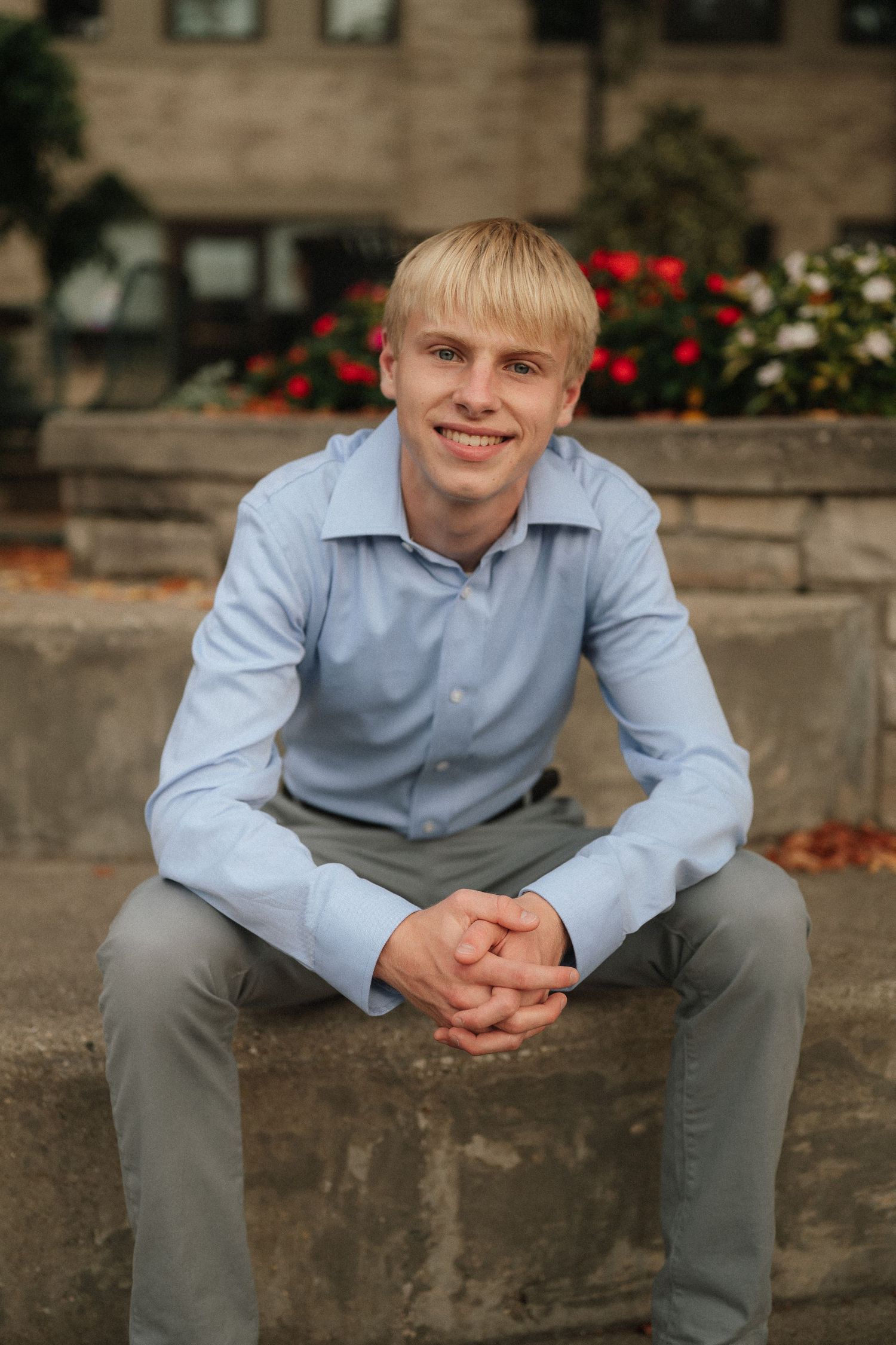 Person in light blue button-down shirt sits casually on stone ledge with red flowers in background.