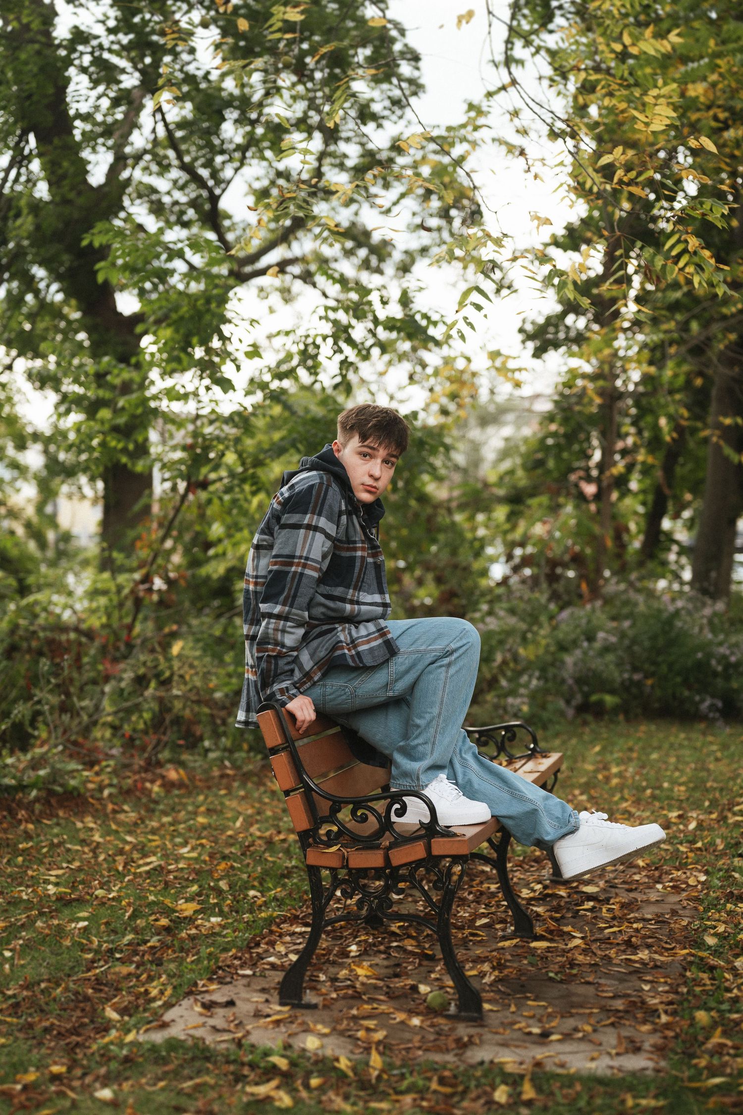 A person in a plaid shirt and jeans sits thoughtfully on a wooden park bench surrounded by autumn leaves and trees.