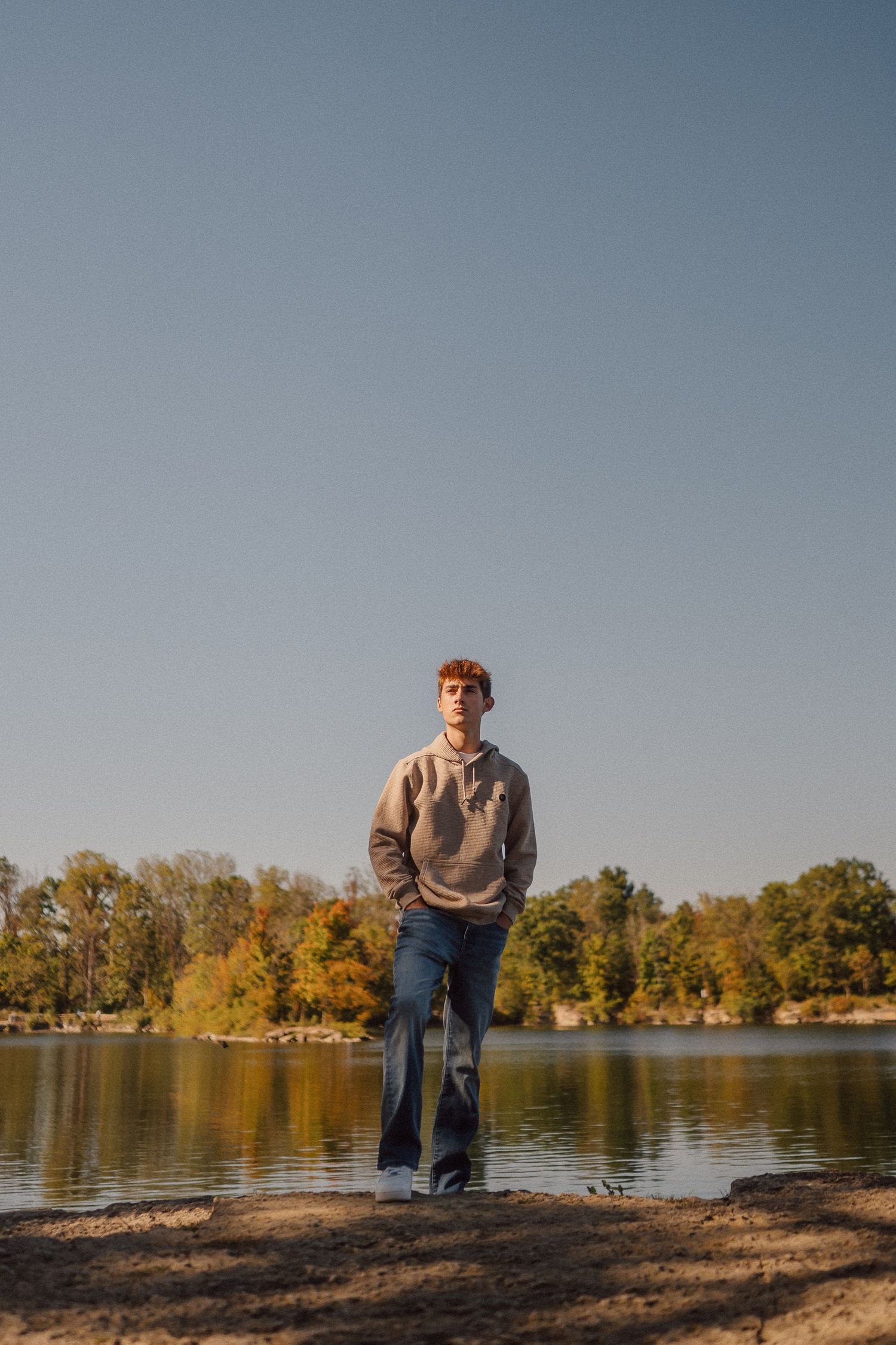 Person posing on rocky shoreline with autumn trees and calm lake waters in the background.