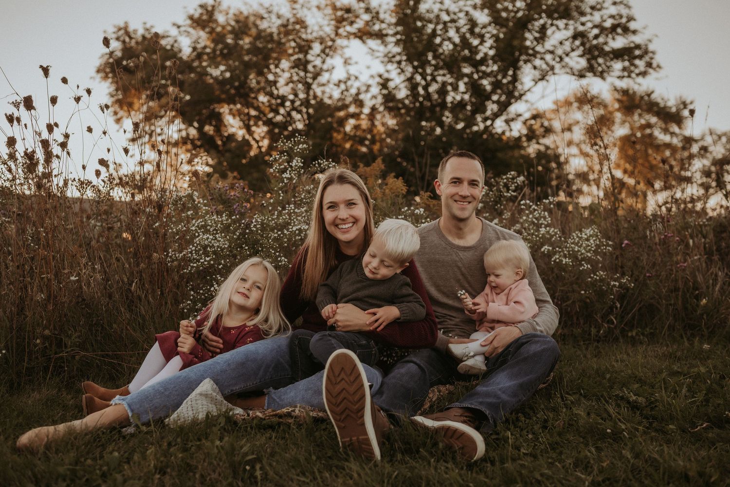 Family relaxing together on grassy field during golden hour sunset with fall foliage in background.