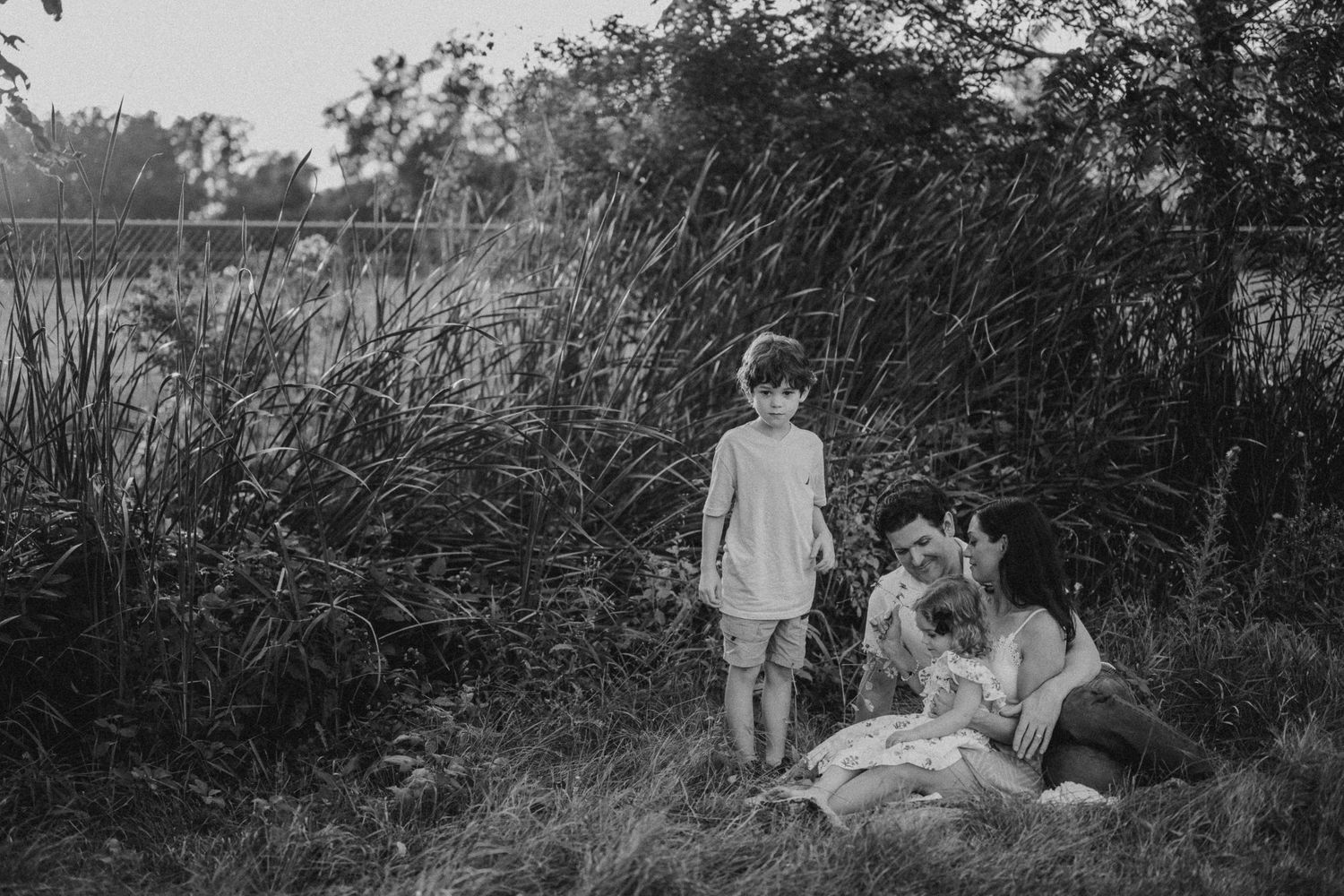 A black and white photo of a family relaxing by tall grass near a lake.