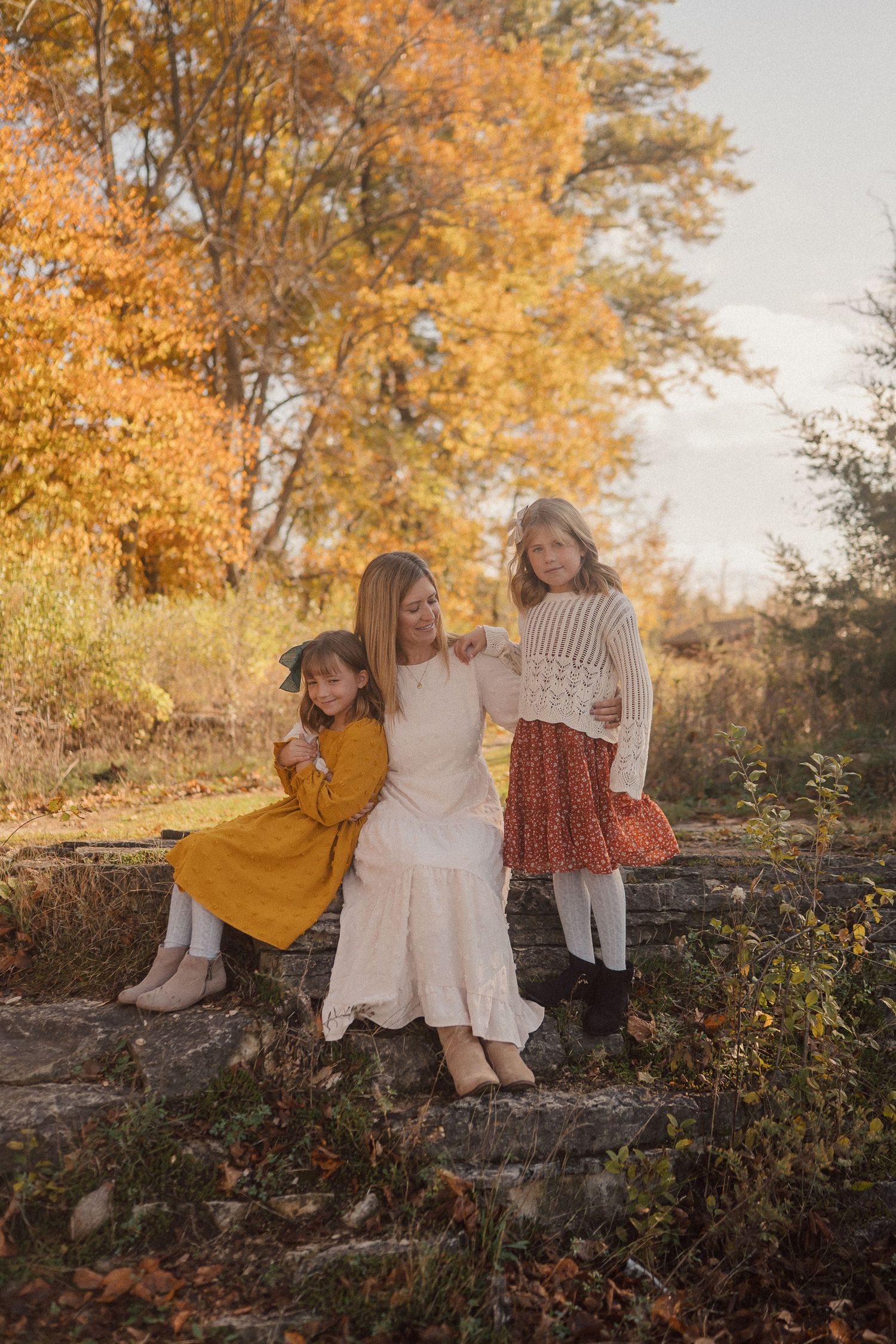 Children sit together on a stone wall surrounded by autumn trees with golden leaves in warm evening light.