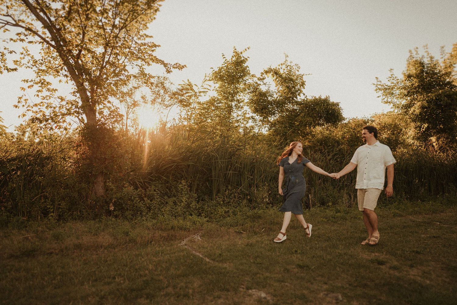 A couple dancing in a grassy field during golden hour with trees in background.