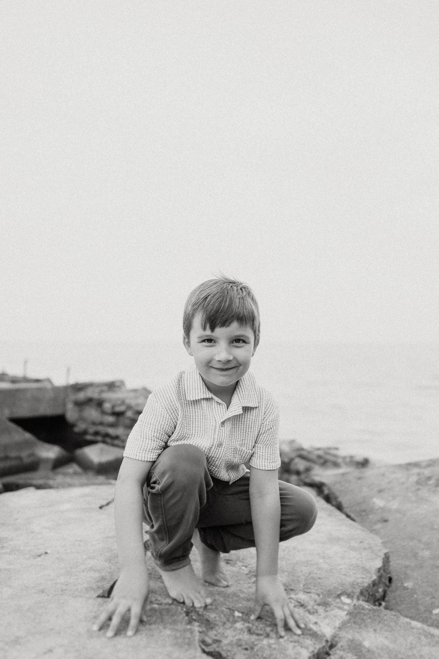 A black and white photo shows someone crouched playfully on coastal rocks by the water.