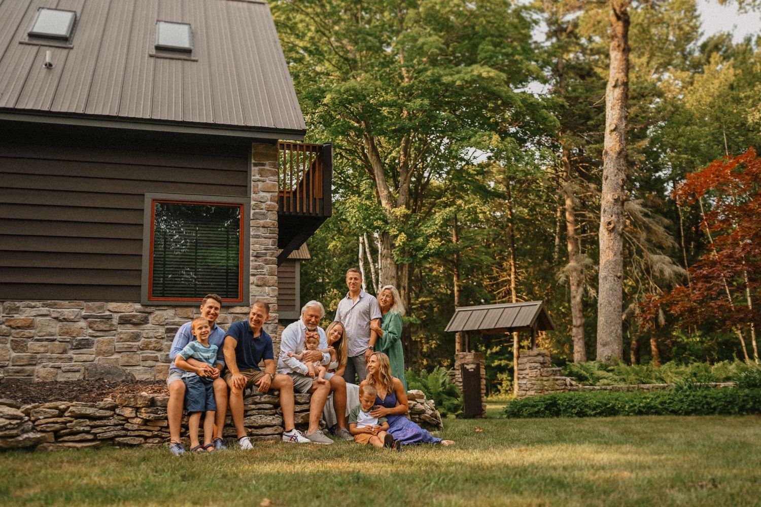 A group gathers on the lawn in front of a modern dark-sided house with stone foundation on a sunny day.