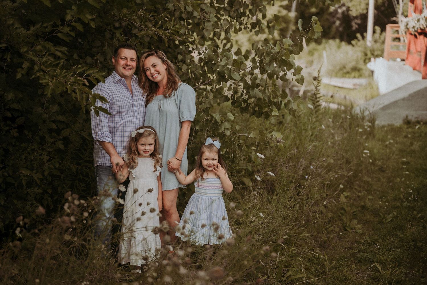 A family of four poses together outdoors in a natural setting with greenery and sunlight in the background.