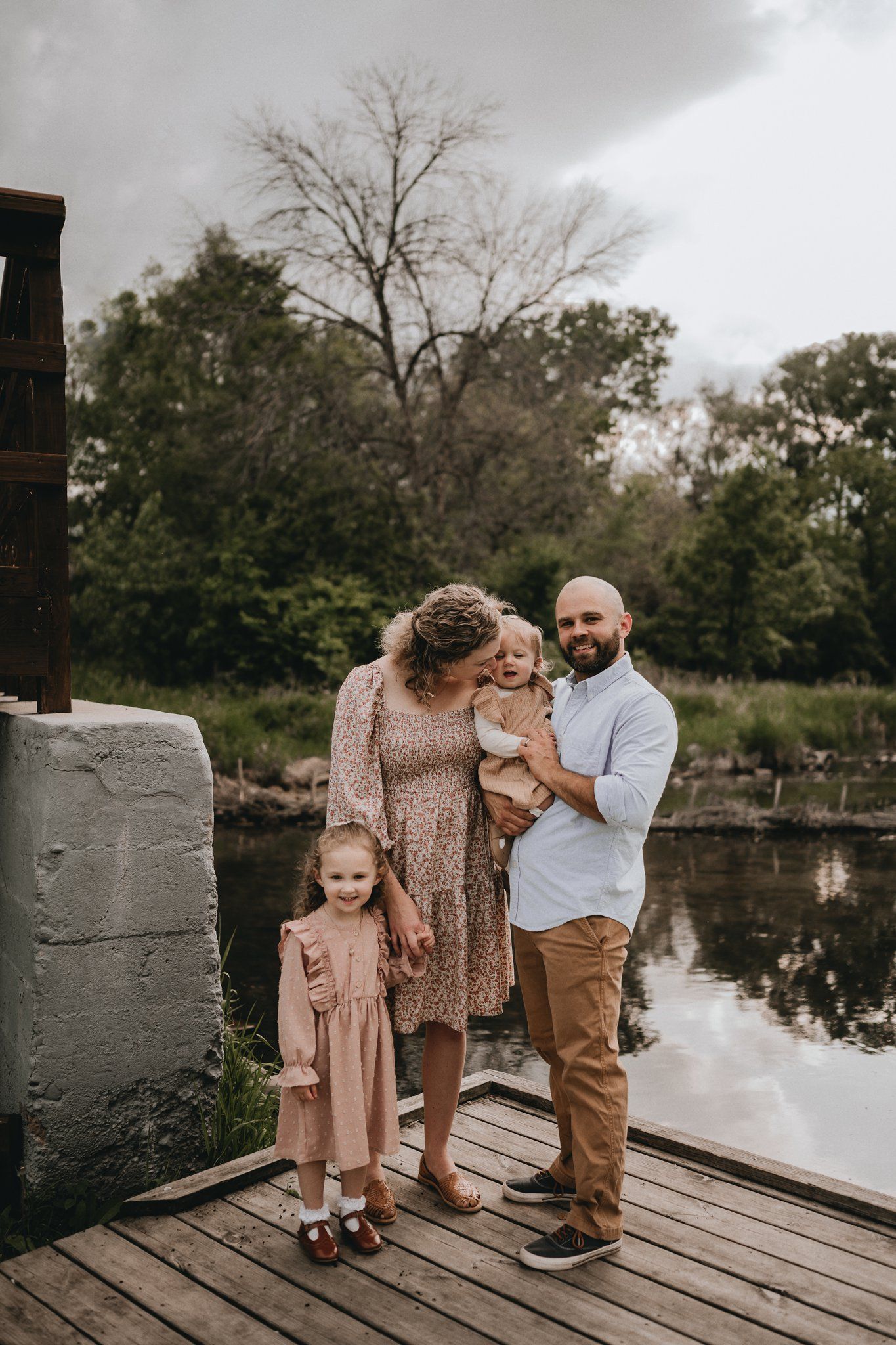 A family portrait on a wooden dock overlooking a serene pond surrounded by trees.