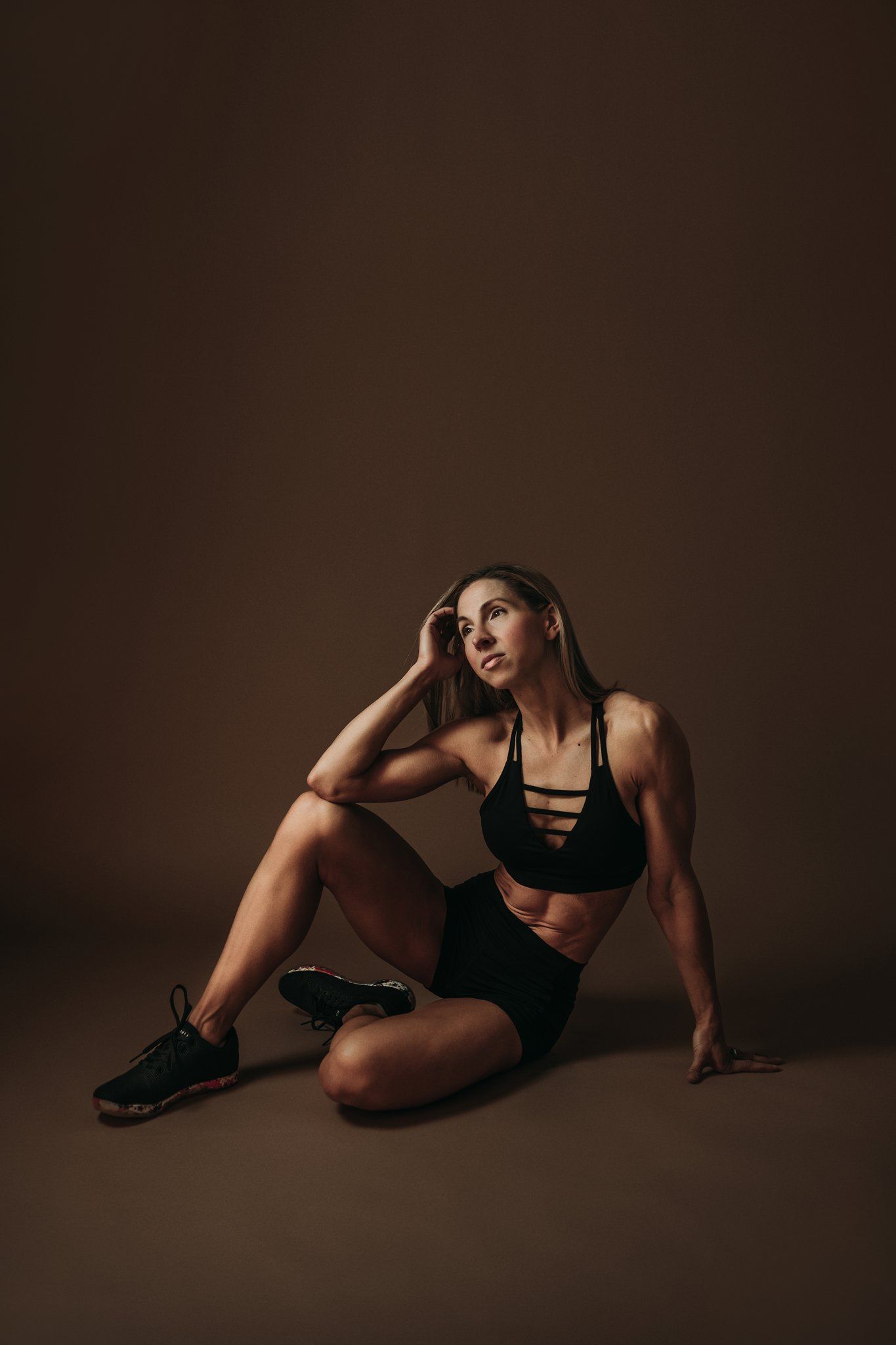 Fitness model in black sportswear sitting on brown backdrop during studio photoshoot.