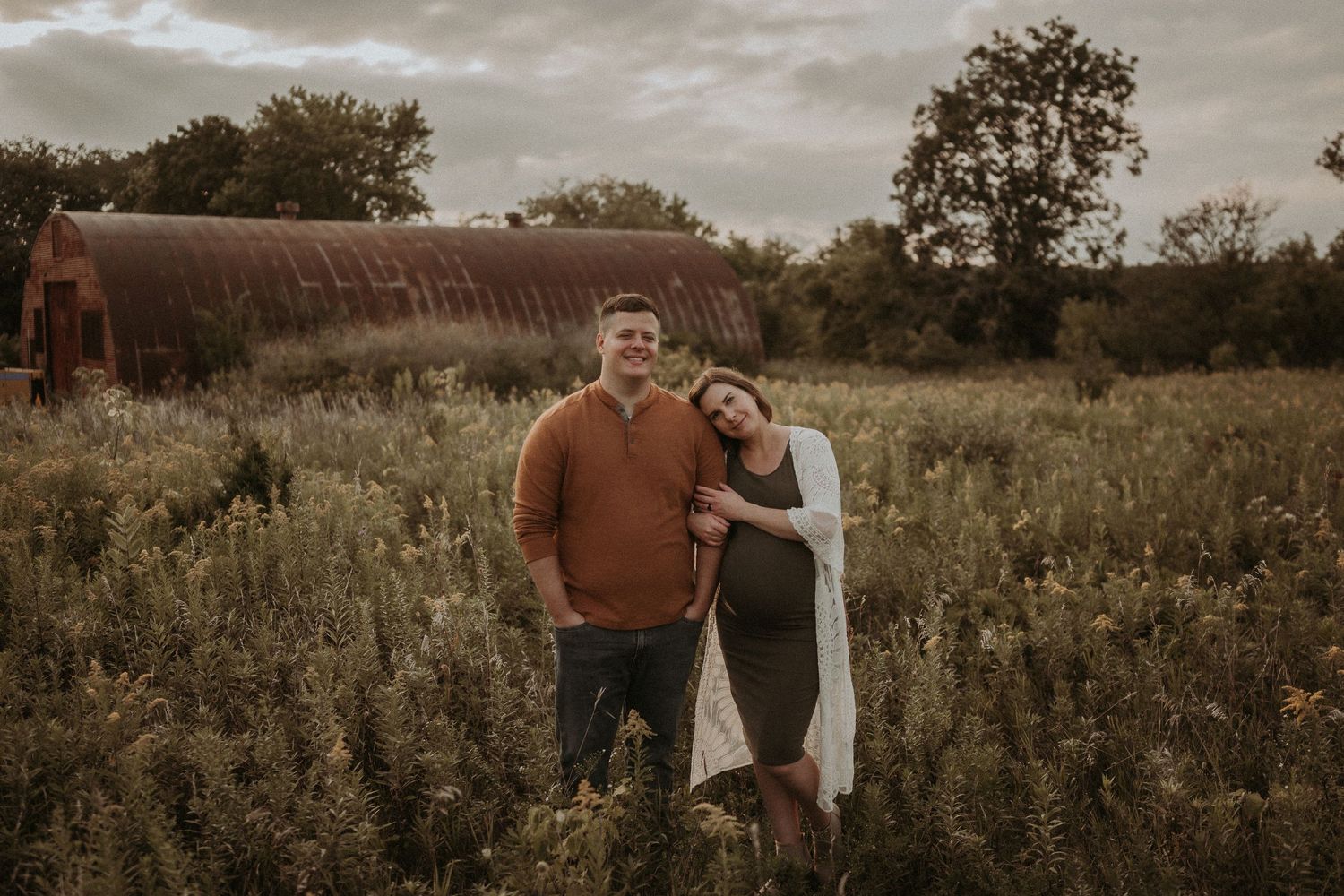Maternity photoshoot in front of rustic barn with couple standing in wildflower field.